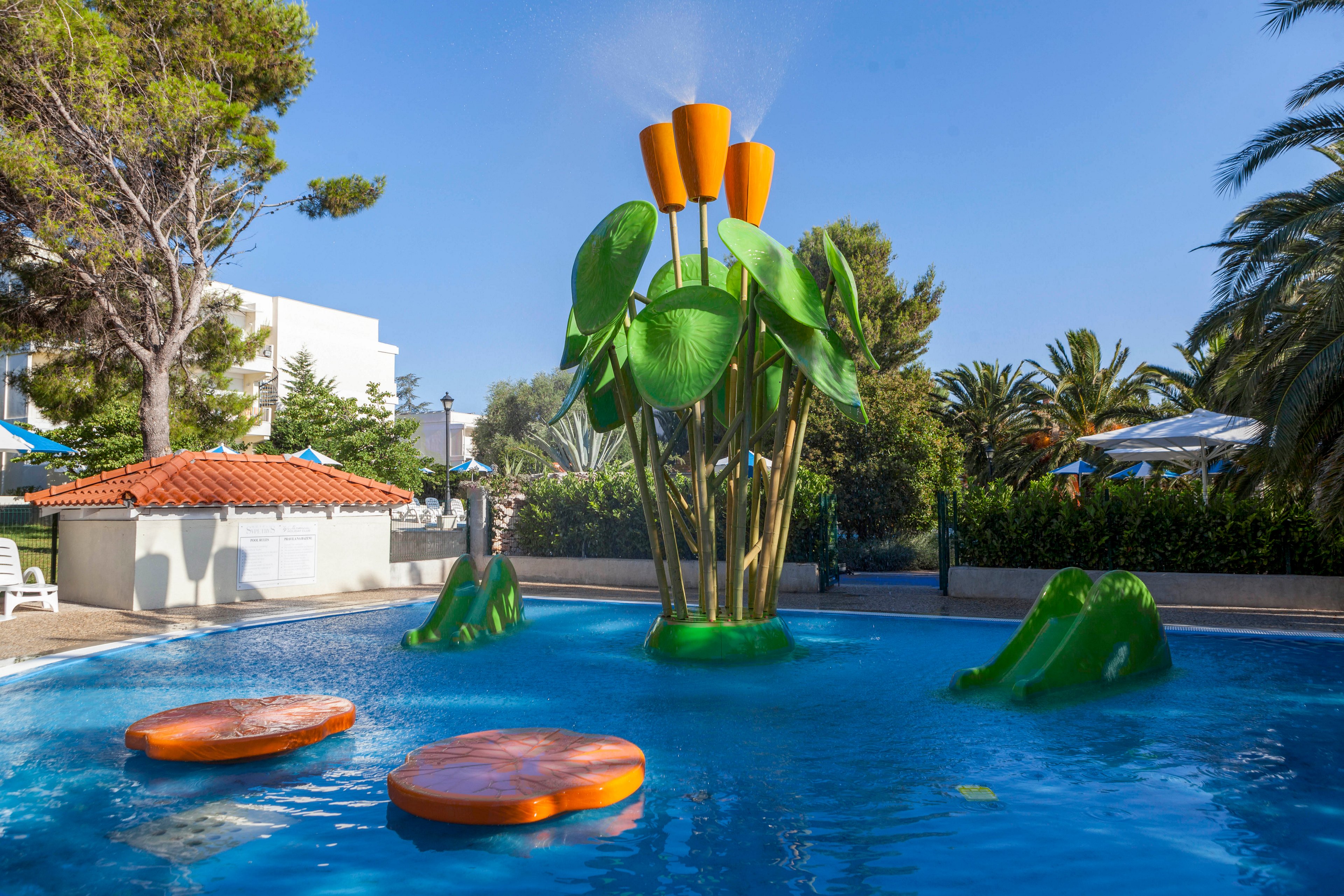 A vibrant outdoor splash pad features a large, whimsical water play structure with oversized green leaves and orange flower shapes spraying water. The pool includes floating circular platforms and small climbing elements, surrounded by lush trees and a sunny blue sky. The setting is playful and inviting, ideal for children’s recreation in a resort or park environment. No visible text or numbers are present in the image.