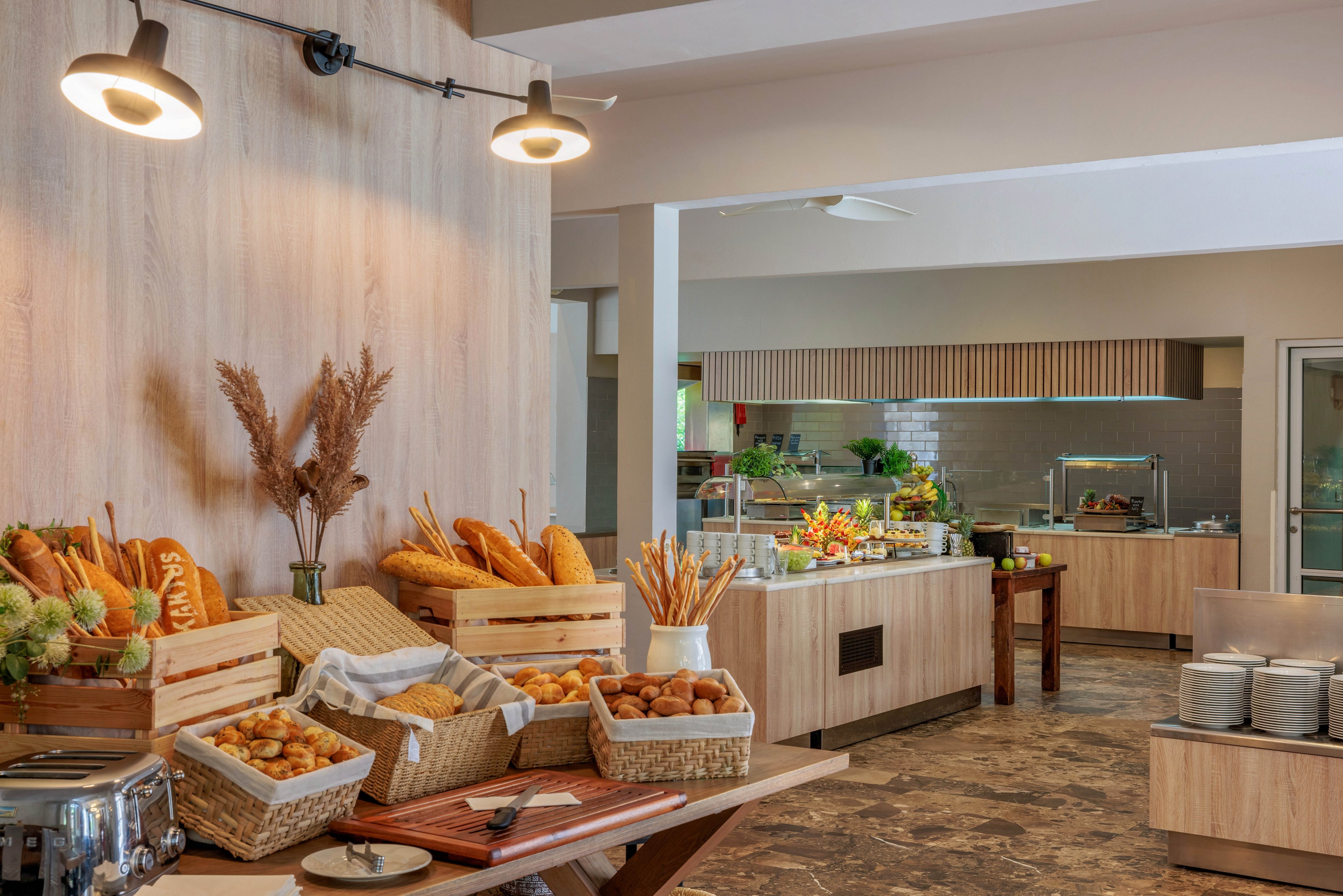 A well-lit breakfast buffet features an assortment of fresh bread, baguettes, and pastries arranged in wicker baskets and wooden crates. The setting is a modern dining area with neutral wood tones and a clean, organized layout. In the background, additional food stations offer fruits and other breakfast items. Decorative dried plants add a rustic touch to the scene. No visible text or numbers are present in the image.