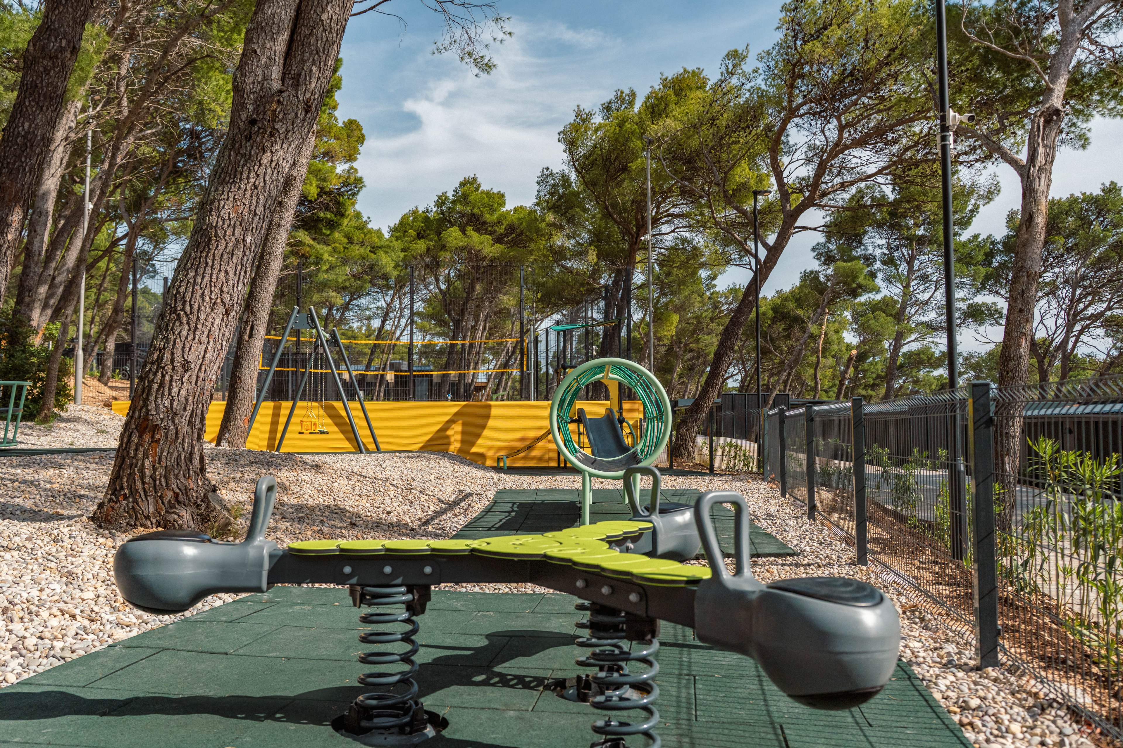 A contemporary spring-mounted seesaw stands in the foreground of an outdoor playground. The play area is set among tall pine trees, with gravel ground cover and rubber safety mats beneath the equipment. In the background, additional play structures and a yellow barrier are visible behind a fence. The scene is brightly lit, suggesting a sunny day in a natural park setting.