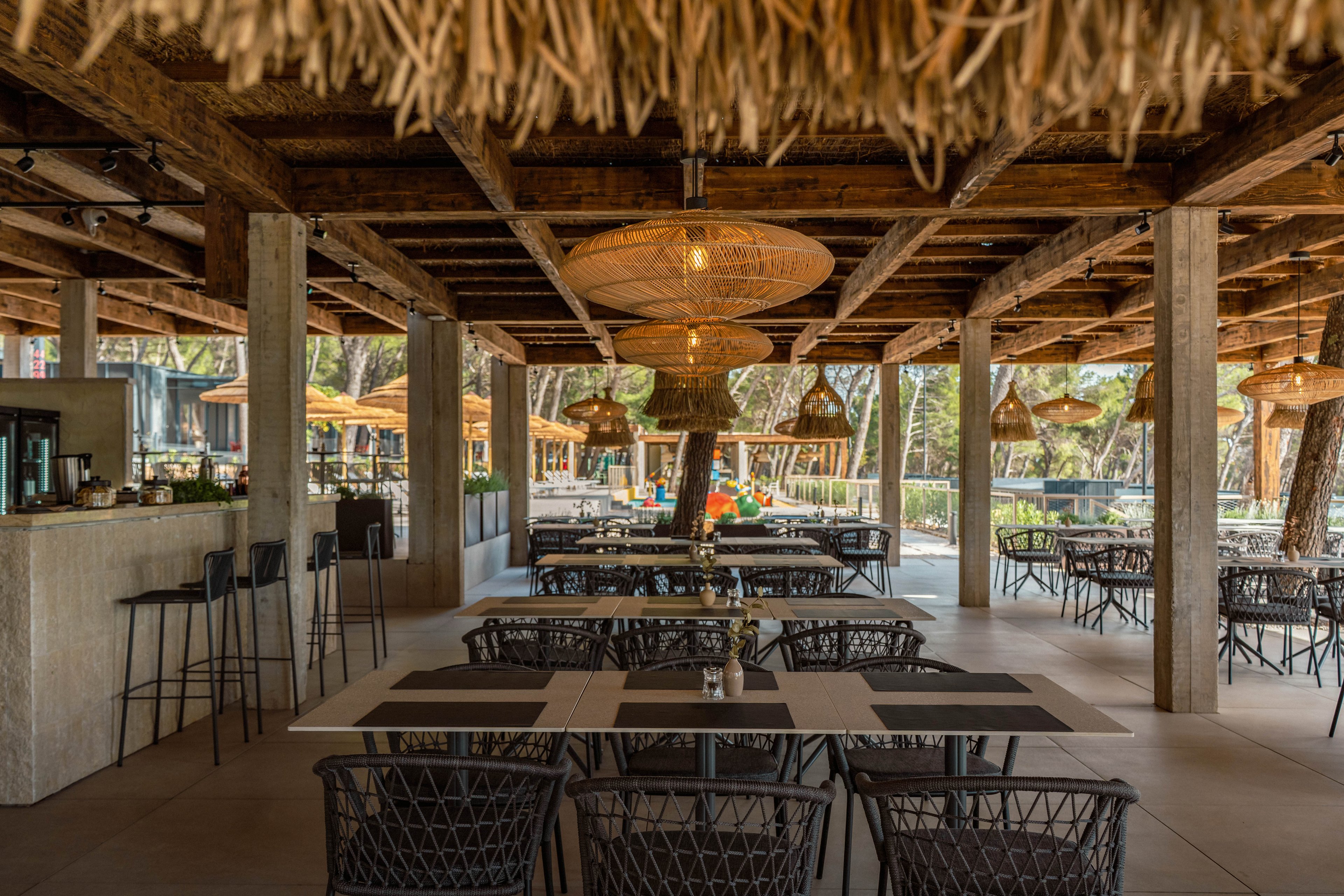 Spacious open-air restaurant interior with wooden beams and thatched ceiling details. Wicker chairs and neatly arranged tables create an inviting dining area facing large windows. Warm pendant lights hang above the seating, enhancing the rustic coastal atmosphere. The bar area with high stools is visible to the left, while trees and outdoor umbrellas can be seen in the background.