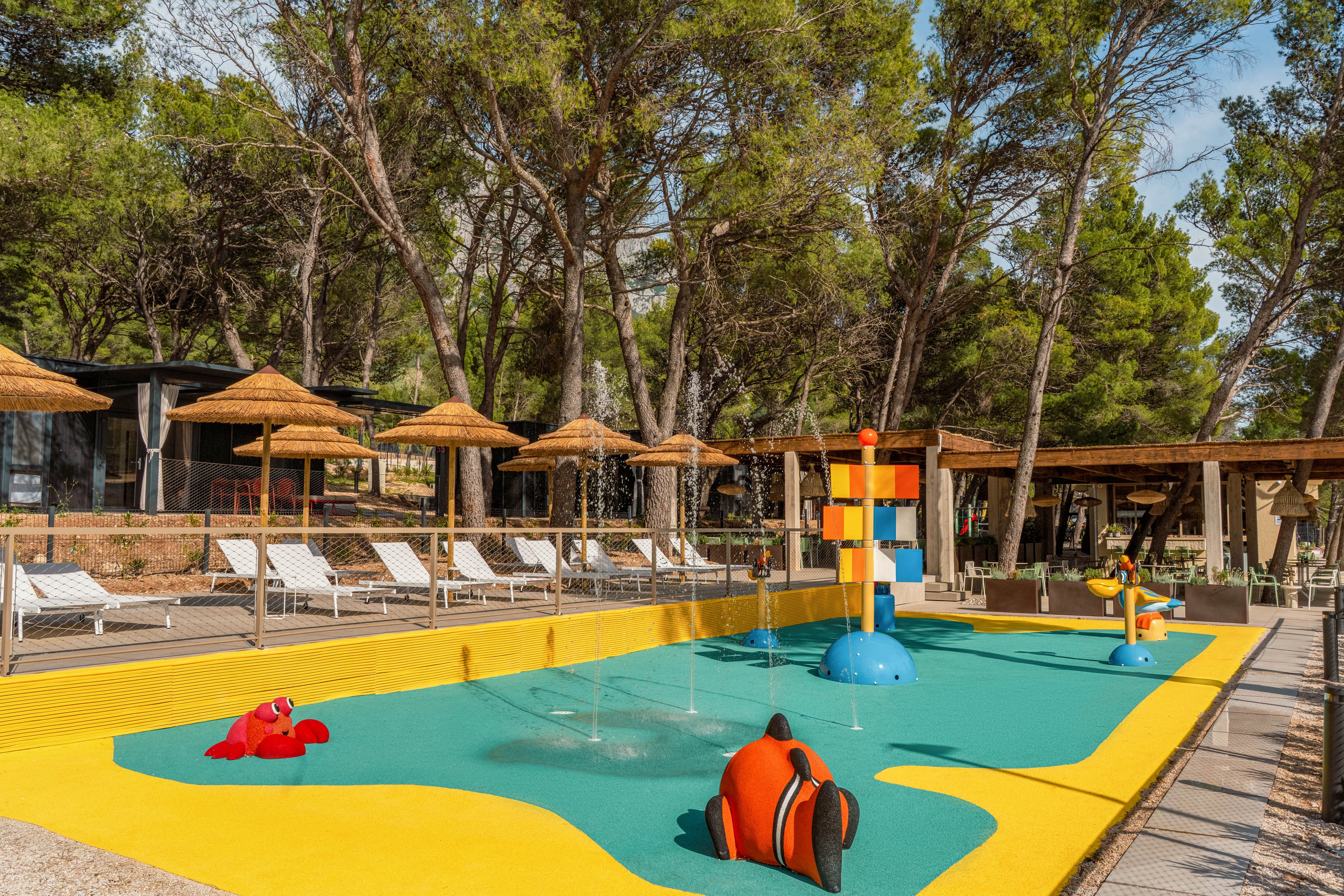A shallow children's splash pool features bright yellow and turquoise flooring with playful sea creature sculptures. Water fountains spray gently from the ground and from a colorful vertical structure in the center. Sun loungers and straw parasols line the deck area around the pool, suggesting a relaxing family-friendly resort. Tall pine trees and wooden structures in the background create a natural, shaded setting.
