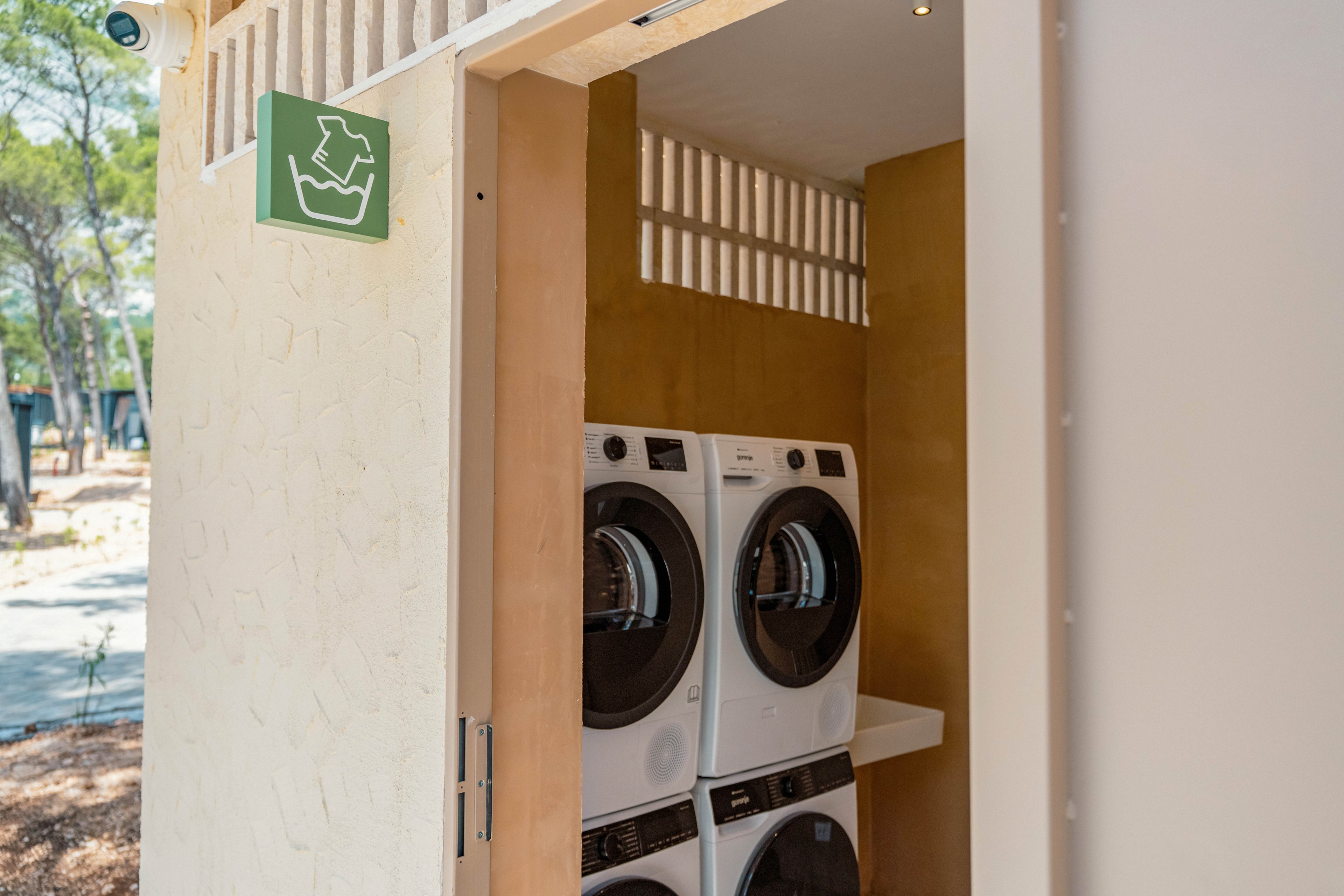 A small laundry room with stacked front-loading washing machines is visible through an open door. The space appears to be part of an outdoor or semi-open facility surrounded by trees. A green sign with a laundry basket icon hangs above the entrance, indicating the washing area. Soft natural light and neutral wall colors create a clean, functional atmosphere.