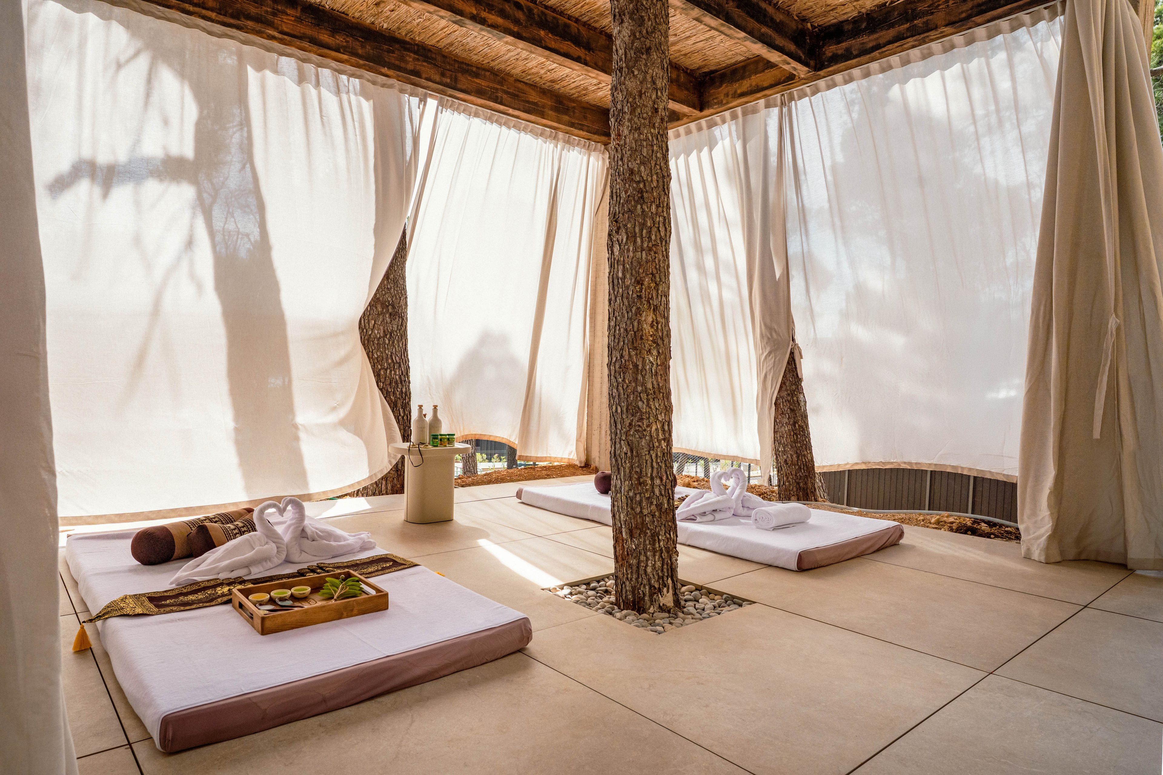An open-air spa cabana features two low massage beds arranged on a tiled floor. Sheer white curtains filter warm sunlight and create a private, tranquil atmosphere around the wooden posts. Rolled towels, a heart-shaped towel arrangement, and a tray with spa essentials are neatly placed on the beds. Bottles and accessories on a small side table enhance the luxurious, relaxing resort setting.