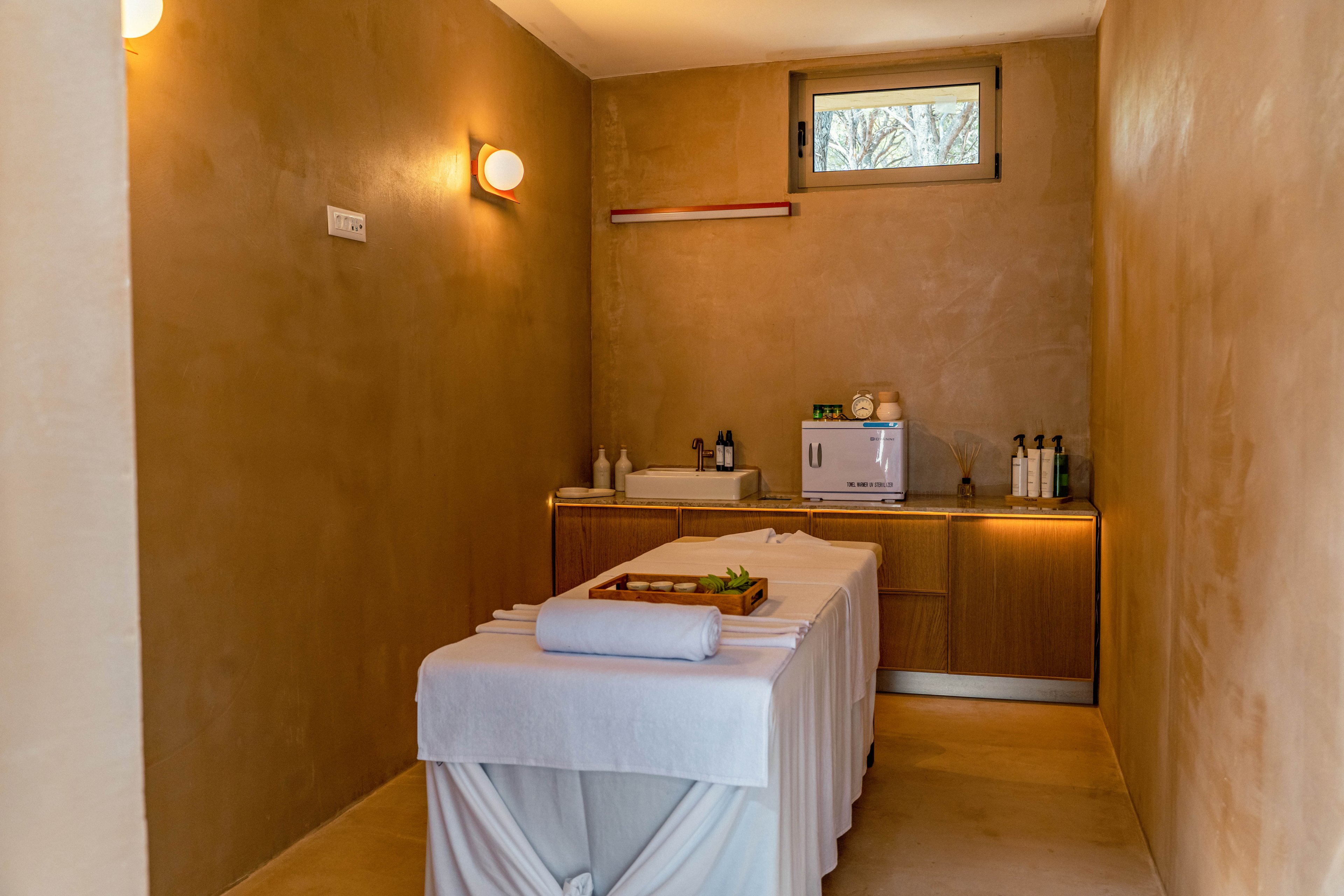 A neatly prepared massage table stands in the center of a minimalist spa treatment room. Warm beige walls and soft lighting create a calm, relaxing atmosphere. Towels are folded on the table with a small tray of green leaves adding a natural touch. At the back, a countertop holds bottles, a small appliance, and other spa essentials beneath a narrow window providing daylight.