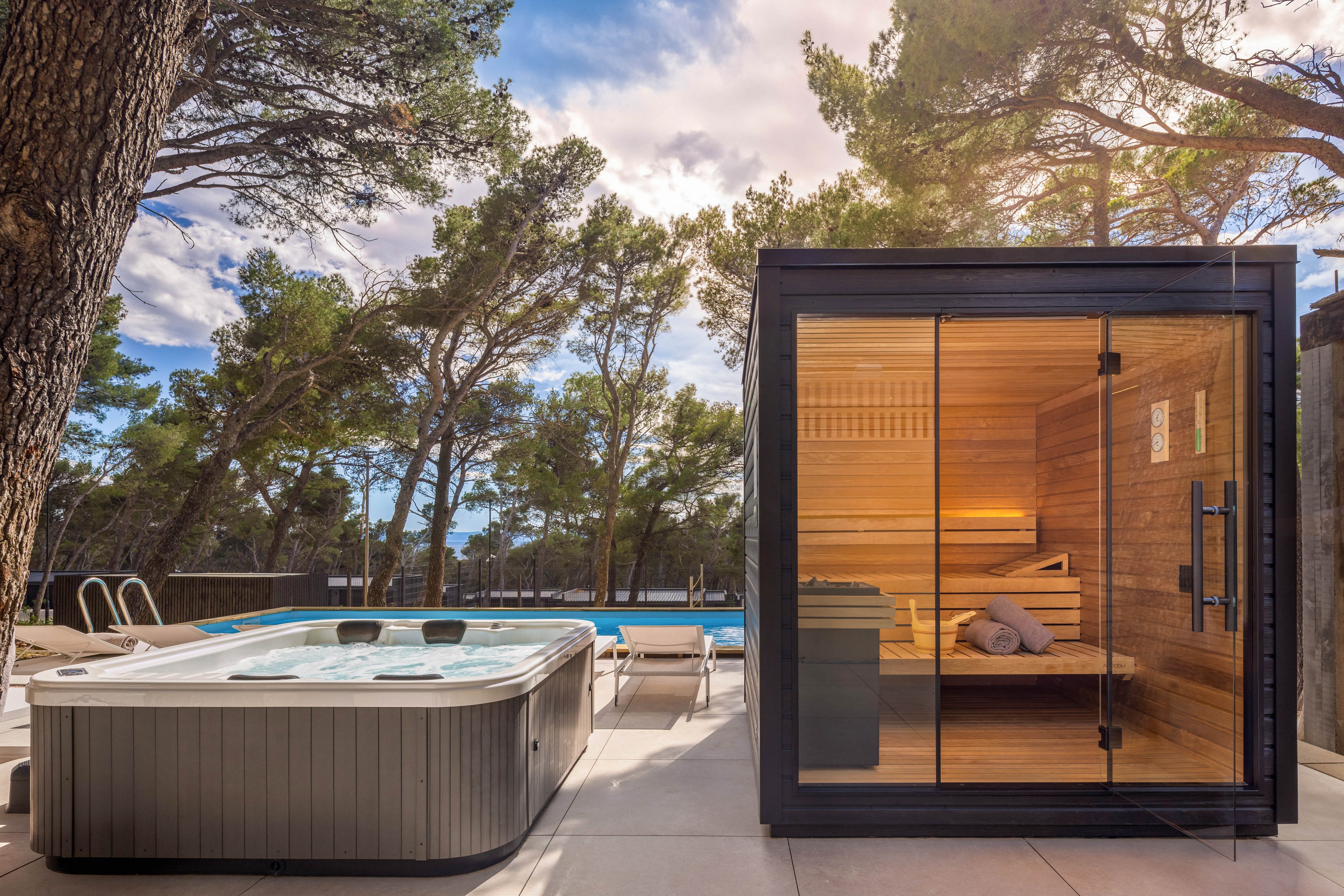A contemporary outdoor wellness area features a glass-front wooden sauna next to a sleek hot tub. The scene is set on a sunlit patio surrounded by tall pine trees and a distant swimming pool. Warm wood tones inside the sauna contrast with the cool blue water and neutral stone flooring. The tranquil environment suggests relaxation, spa treatments, and luxury vacation living.