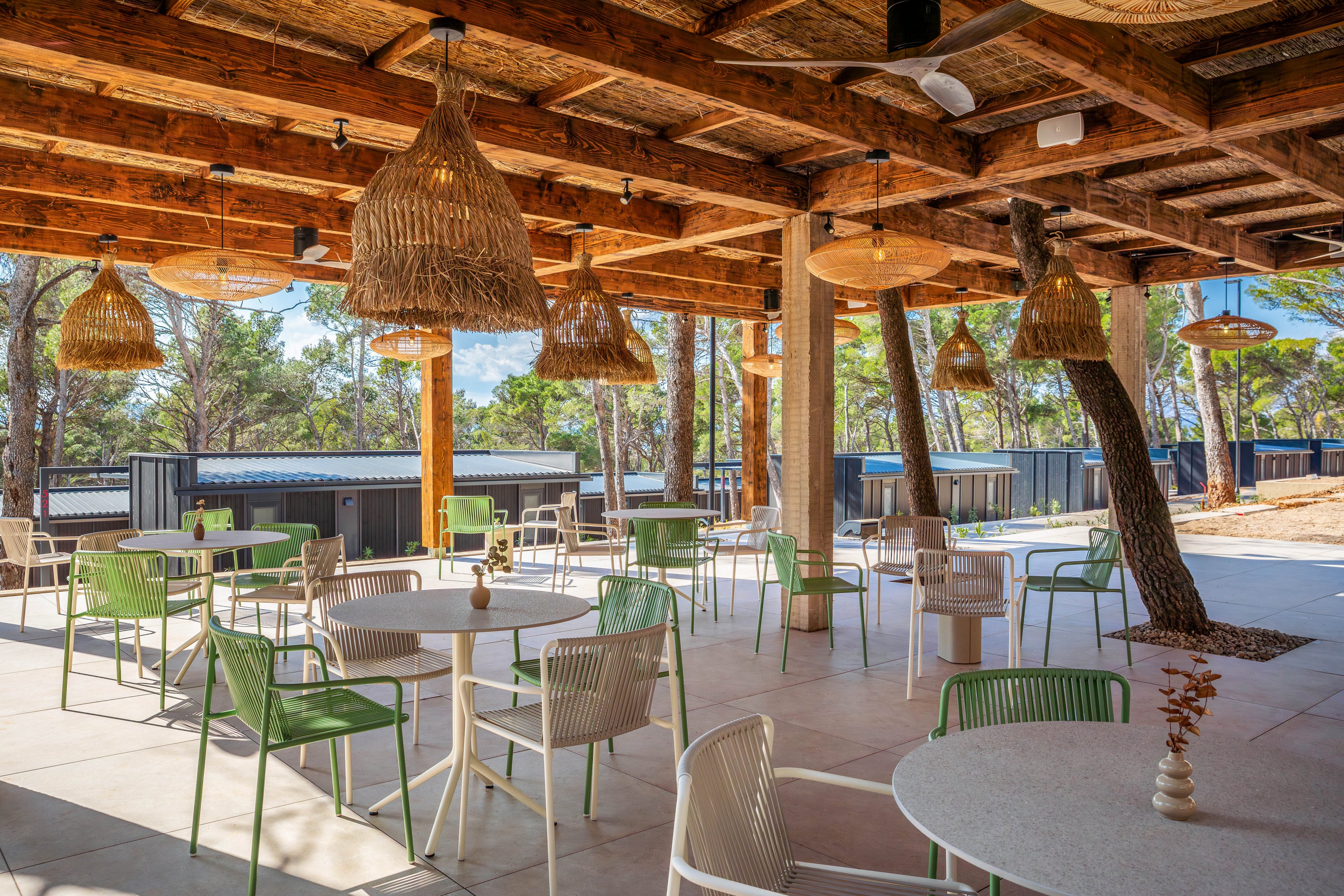 An open-air café terrace features round tables and modern chairs arranged beneath a rustic wooden pergola. Woven rattan pendant lights hang from the ceiling, casting warm tones over the tiled floor. The seating area is surrounded by trees and natural greenery, creating a relaxed outdoor atmosphere. Soft natural light and earthy materials emphasize a casual, vacation-like setting.
