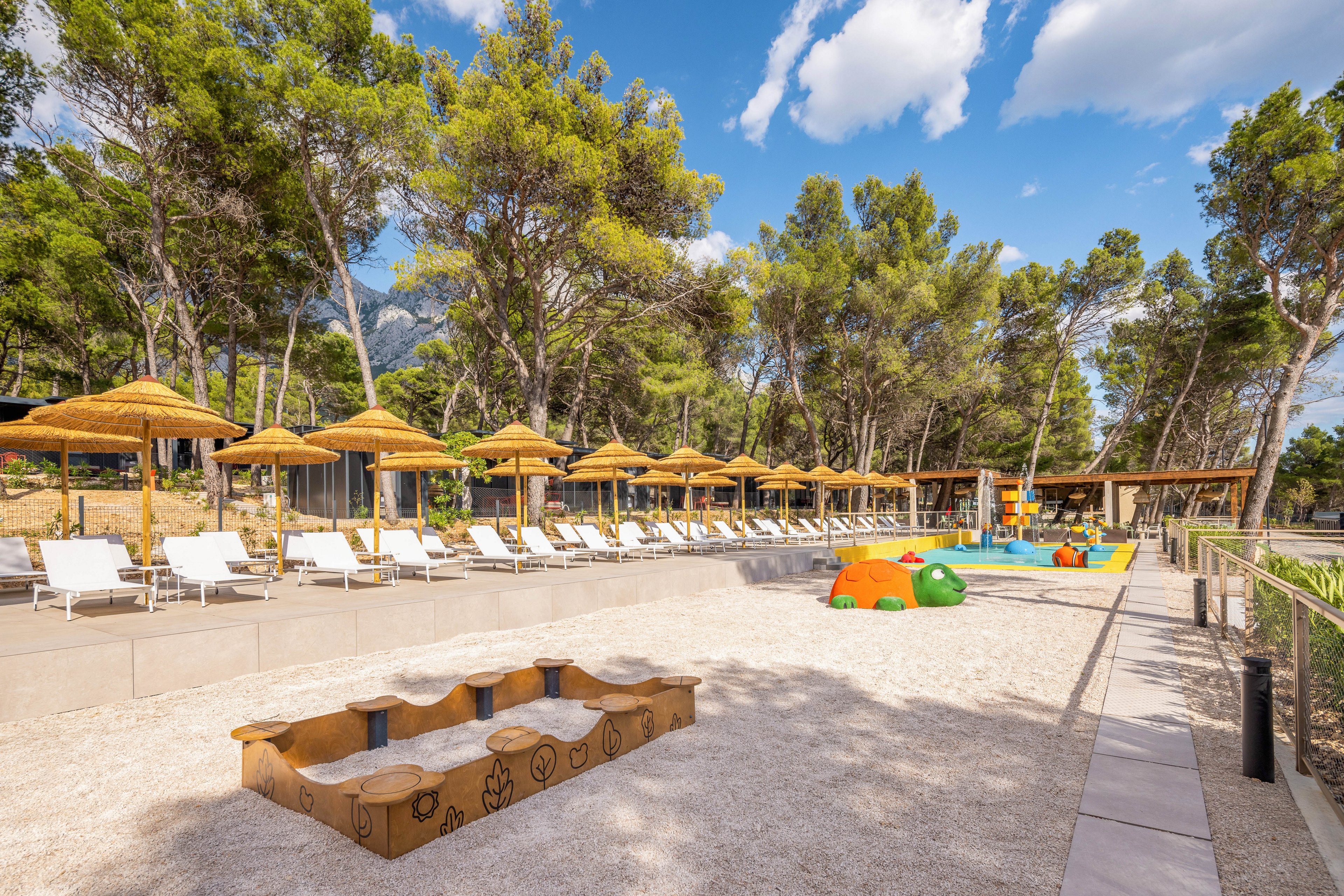 Outdoor resort area featuring rows of white sun loungers under straw parasols is shown on a bright sunny day. In the foreground, a sandy play zone with a low wooden bench decorated with simple drawings faces the pool area. Colorful animal-shaped play structures and a small splash zone are visible near the pool. Tall green pine trees and blue sky with scattered clouds create a relaxing holiday atmosphere.