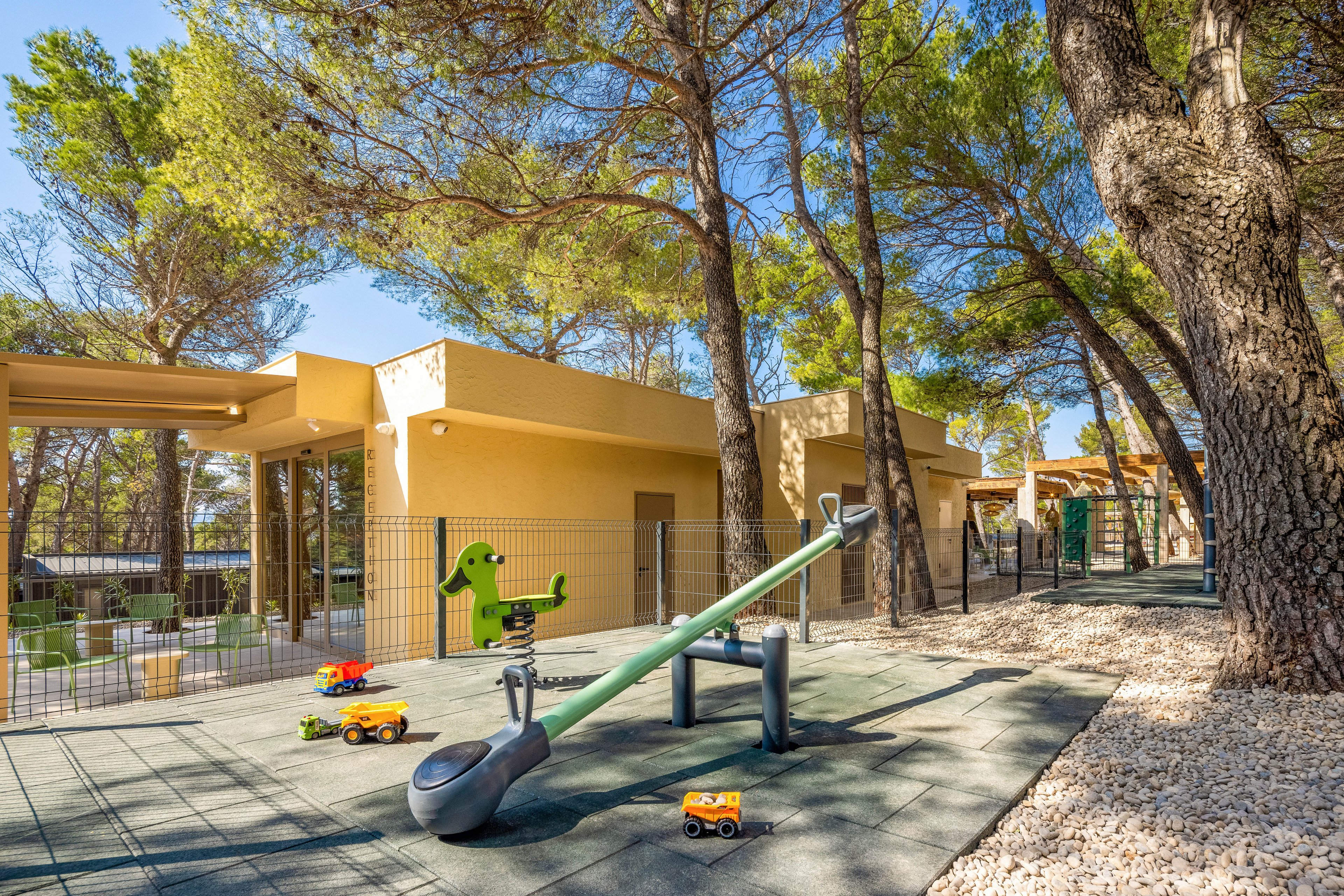 An outdoor playground features a modern green seesaw and a spring rider surrounded by tall pine trees. Small toy trucks are scattered on the rubber safety flooring, adding bright color accents. A low fence encloses the play area, with a contemporary yellow building in the background. The scene is sunlit and inviting, suggesting a safe and cheerful space for children to play.