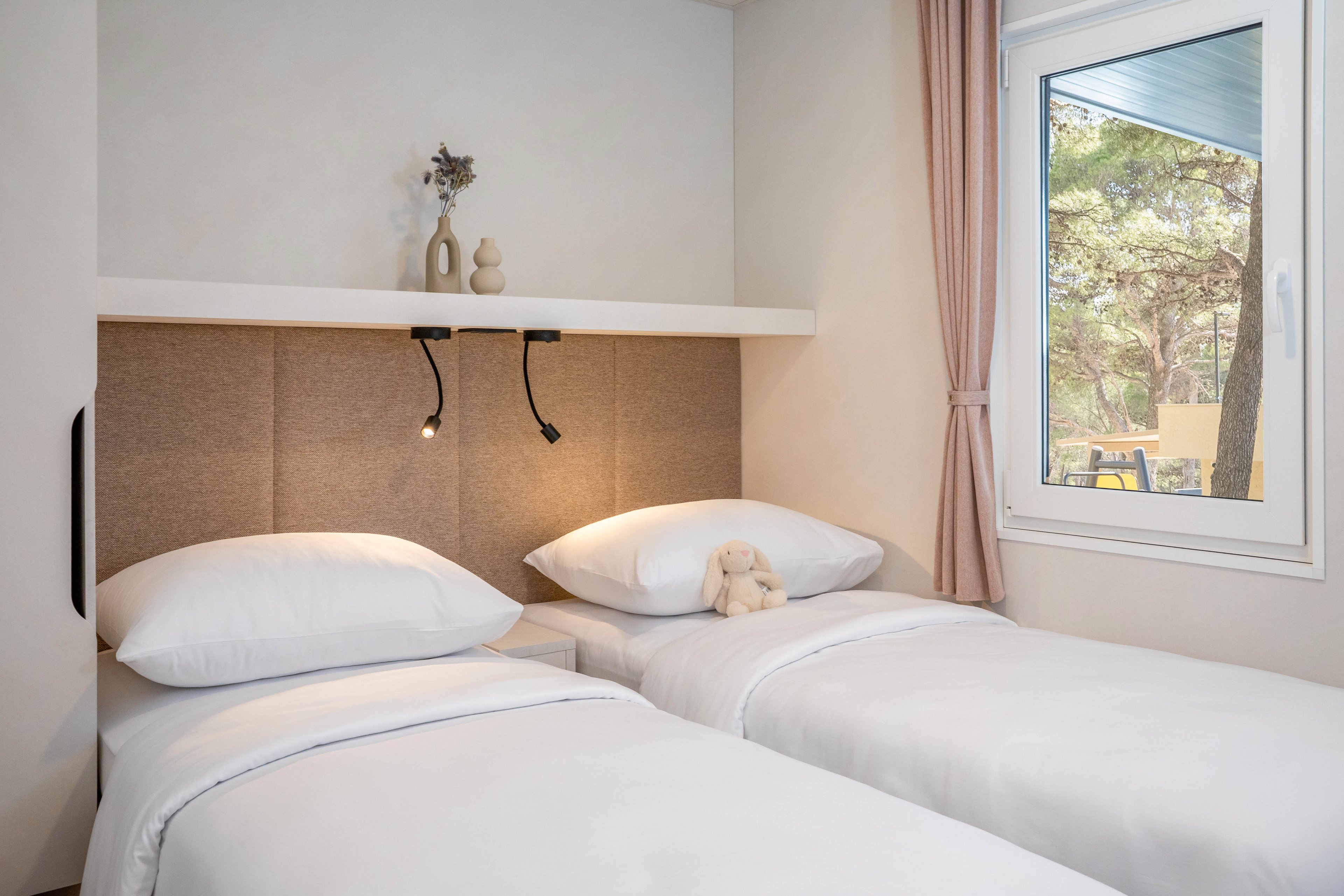A minimal twin bedroom features two neatly made single beds with white linens. A small plush toy rests on one of the beds, adding a soft, welcoming touch. Above the headboards, a shelf holds simple decorative vases and two flexible reading lights. A large window with pink curtains reveals trees and outdoor seating, bringing natural light into the calm, neutral-toned room.
