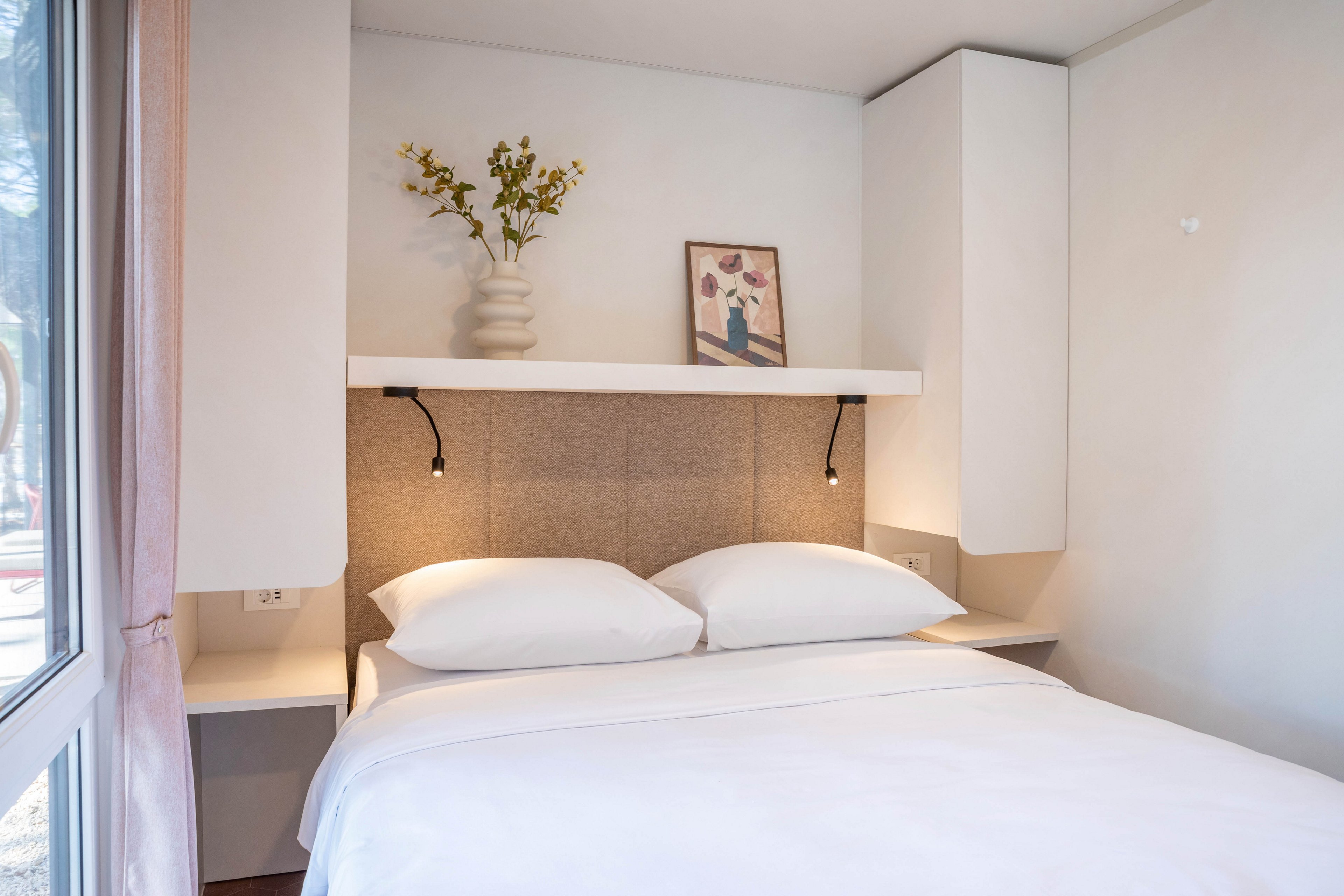 A neatly made bed with white linens is the focal point of this minimalist bedroom. Built-in shelves above the headboard hold a vase of flowers and a framed artwork, adding subtle decoration. Soft wall-mounted reading lights create a warm and inviting atmosphere. Large windows on the side allow natural light to brighten the clean, modern space.