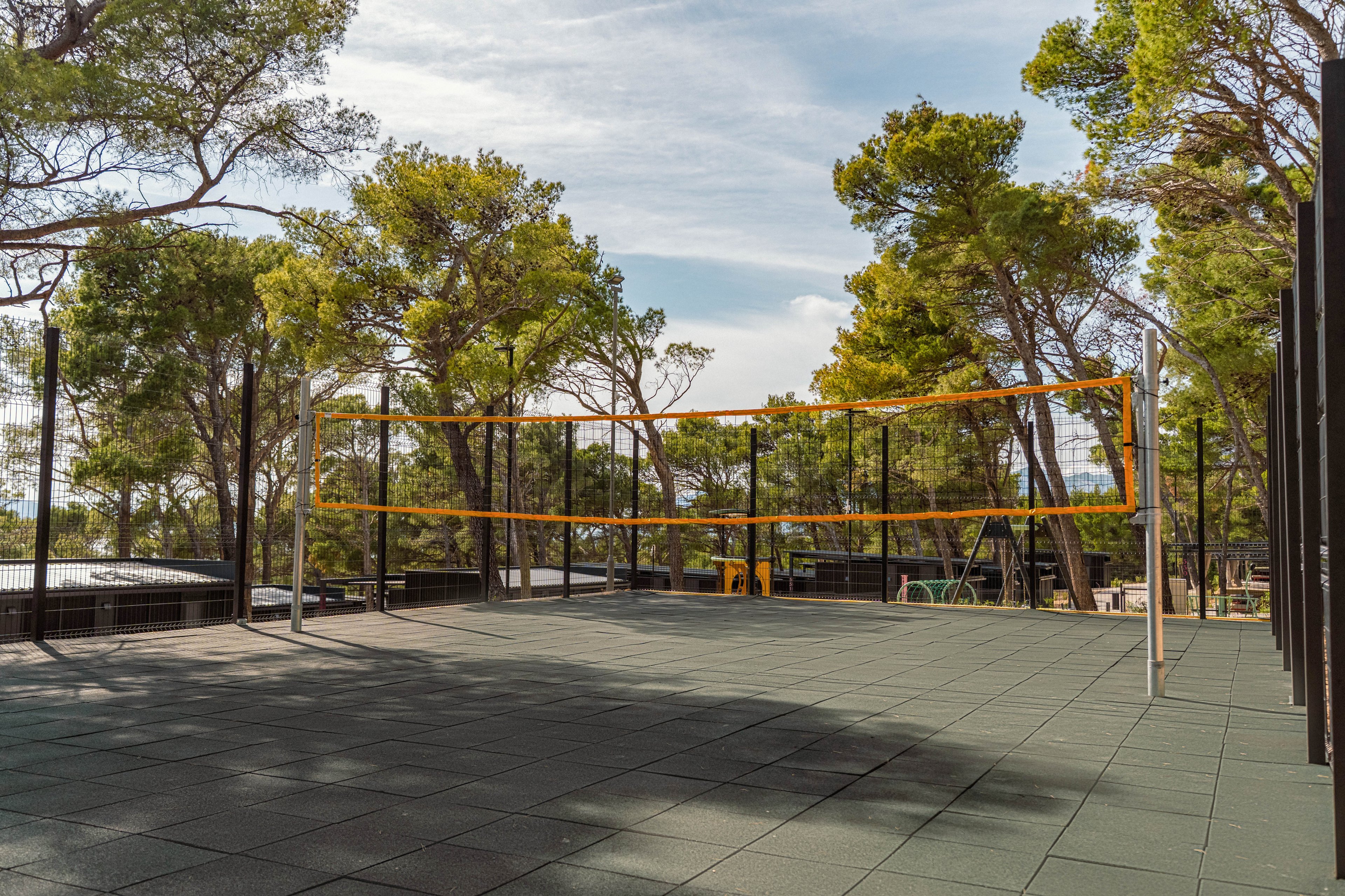 An empty outdoor volleyball court is set up with a yellow net stretched between metal poles. The court surface is made of dark rubber tiles and is enclosed by a tall fence. Pine trees and natural greenery surround the area, with a glimpse of the sky and distant landscape in the background. The scene feels calm and ready for recreational sports or leisure activities.
