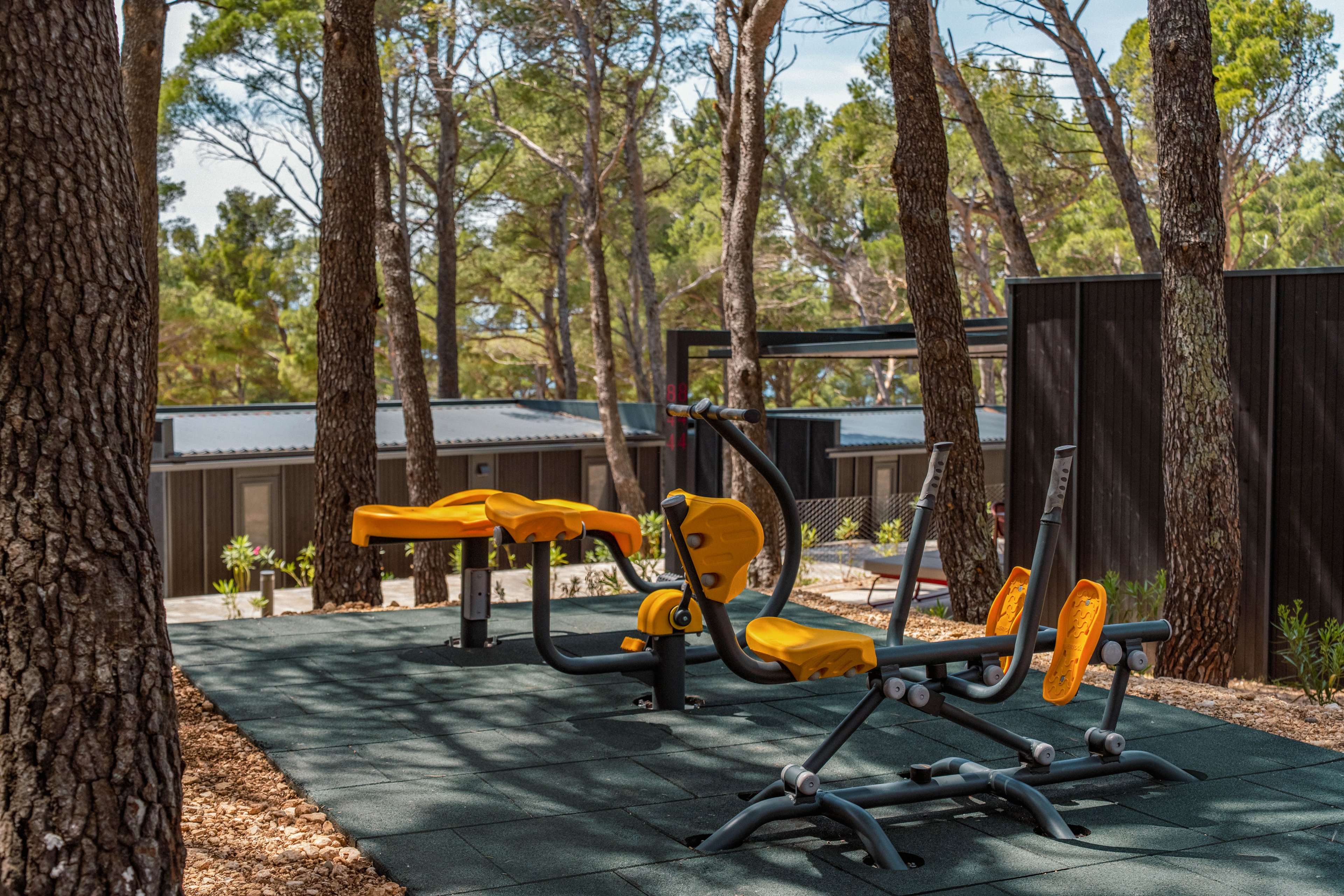 Bright yellow outdoor gym machines stand on a rubberized exercise surface in a wooded area. Tall pine trees surround the fitness zone, creating a natural, shaded environment. Modern dark-colored buildings are visible in the background, suggesting a resort or campground setting. The scene is quiet and empty, emphasizing the peaceful atmosphere of exercising in nature.