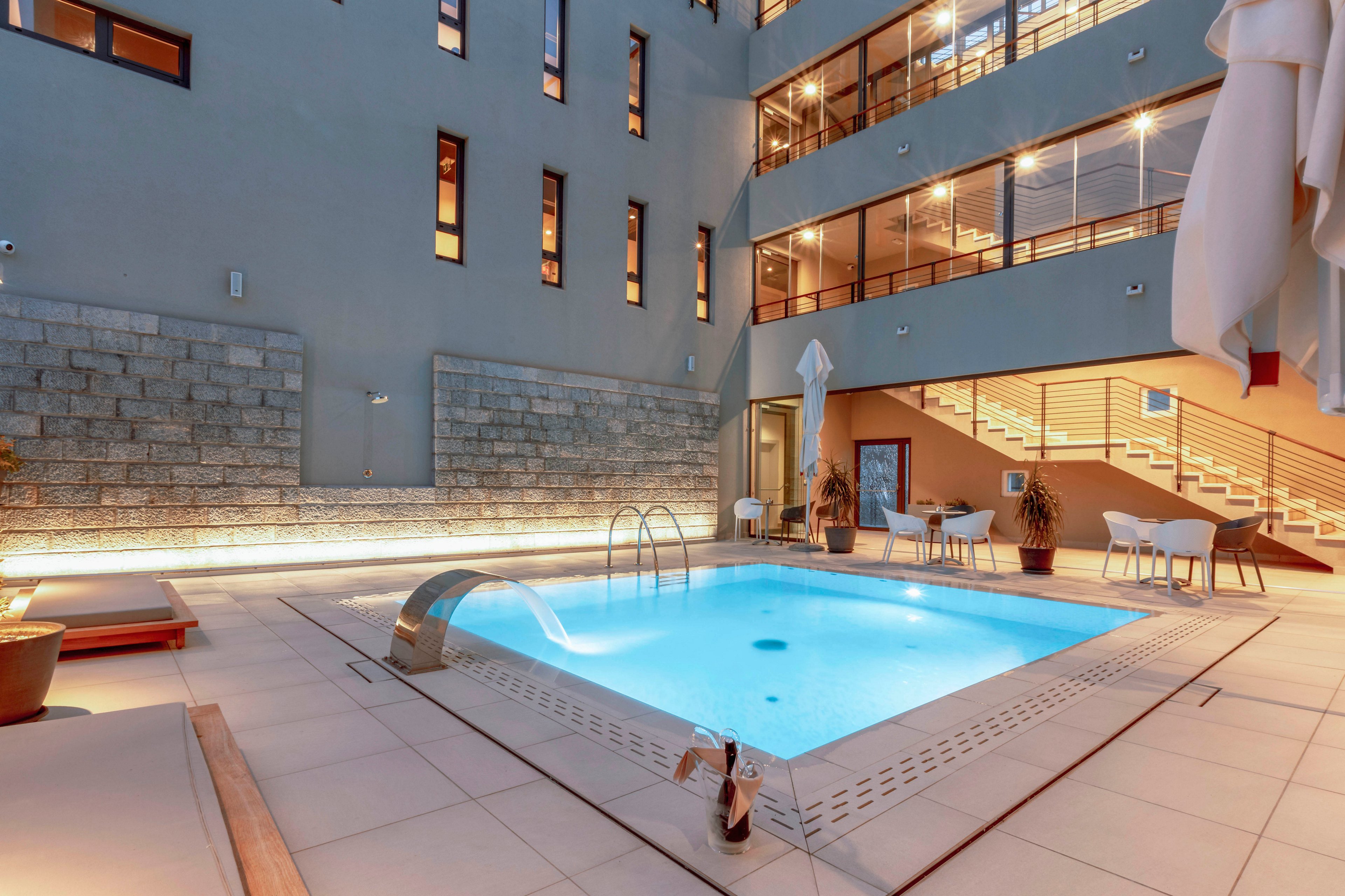 A contemporary hotel courtyard features a rectangular swimming pool surrounded by lounge chairs and tables. The setting is illuminated with warm lighting, highlighting the clean architectural lines and glass windows of the building. The pool area is empty, creating a tranquil and inviting atmosphere. Neutral tones dominate the palette, with touches of blue from the water and soft beige from the tiles. No visible text or numbers are present in the image.