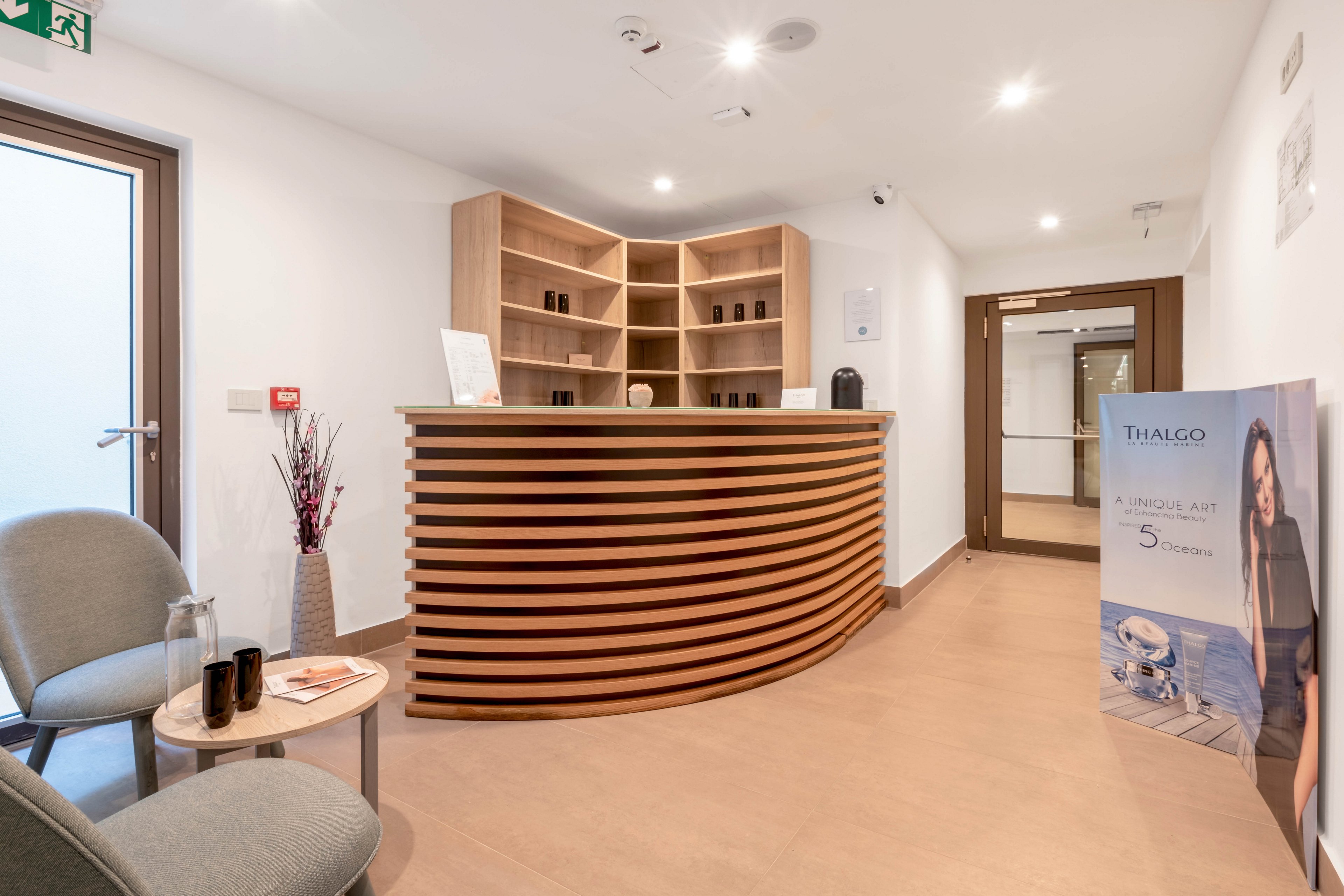 A contemporary spa reception area features a curved wooden desk with minimalist shelving behind it. The setting includes two gray chairs and a small table with cups, creating a welcoming atmosphere. A visible sign displays 'THALGO' and the number '5', suggesting a spa or wellness promotion. The space is well-lit with neutral tones and clean lines, evoking a calm and professional mood.