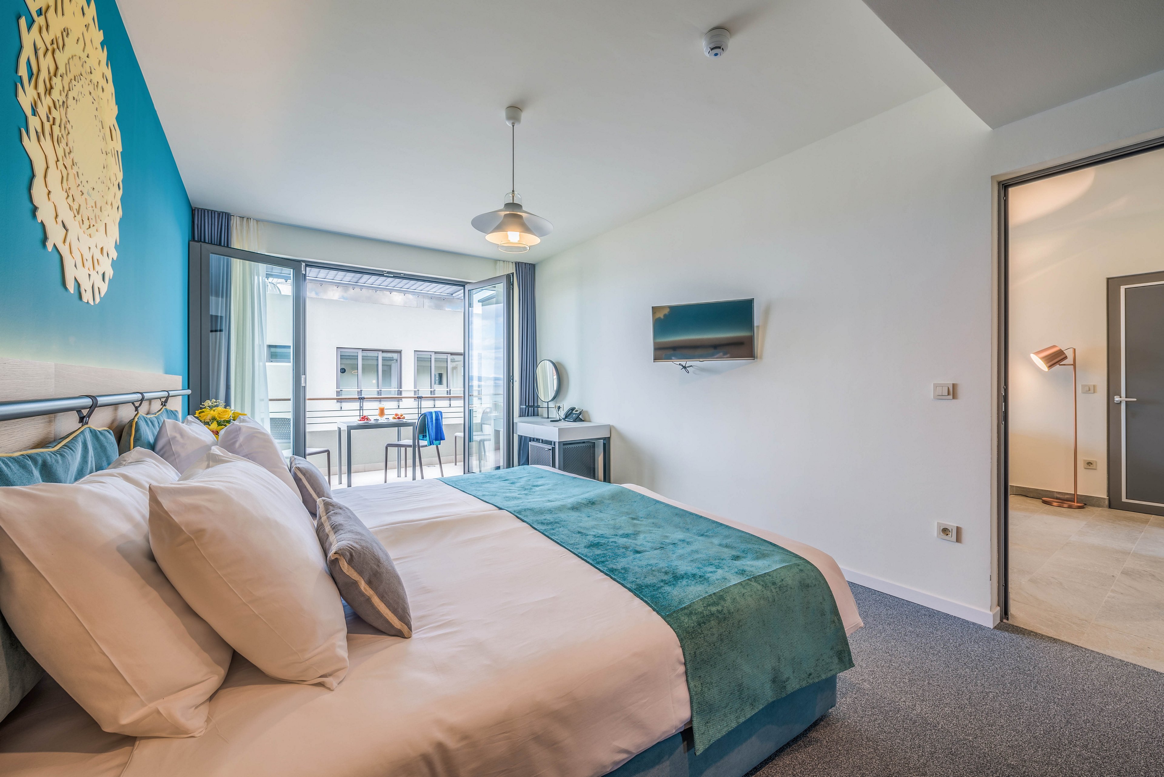 A contemporary hotel room features a large bed with multiple pillows and teal bedding. The room opens onto a balcony with outdoor seating, visible through wide glass doors. The interior is decorated with a blue accent wall, wall art, and a mounted television. Natural light fills the space, creating a bright and inviting atmosphere. No visible text or numbers are present in the image.