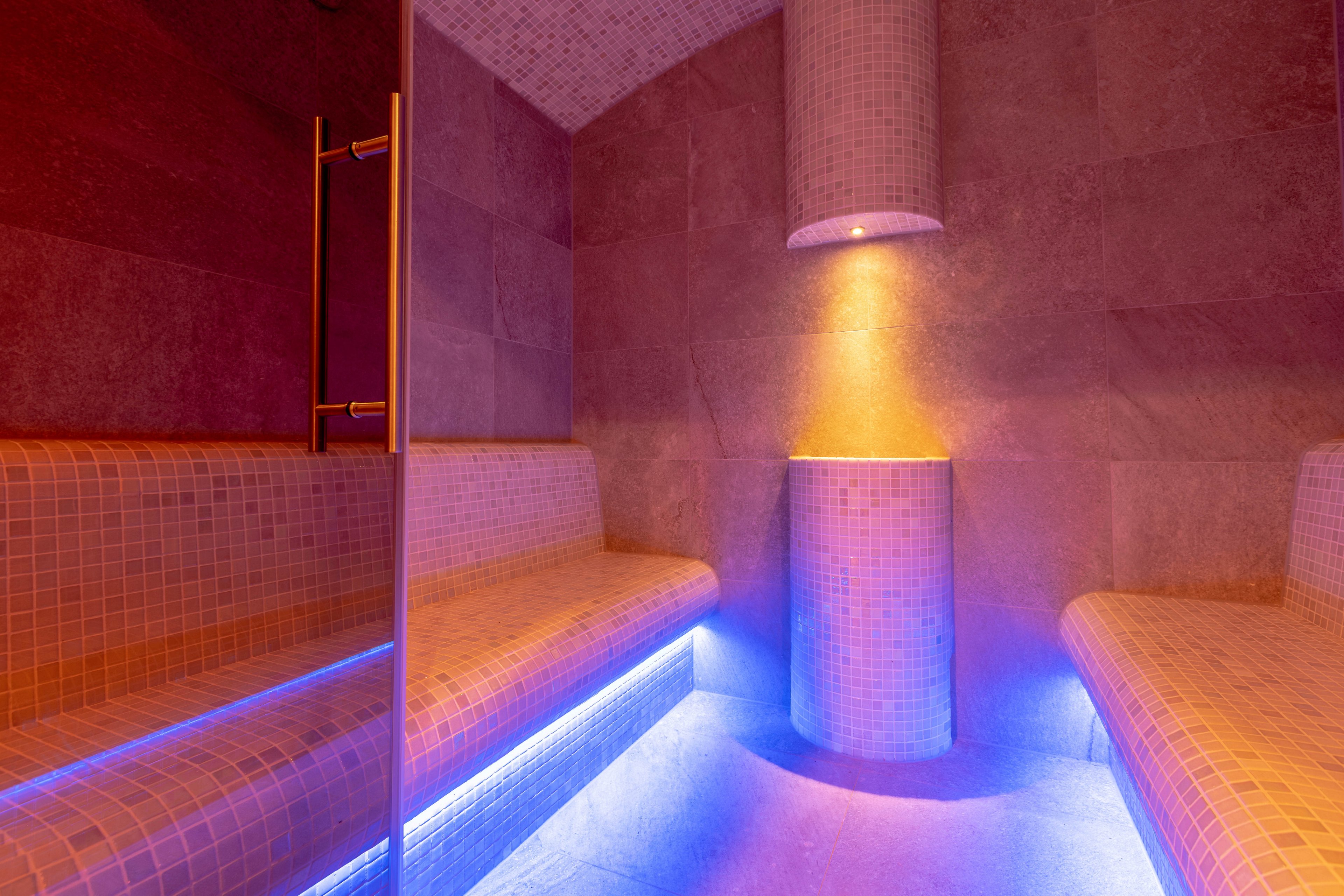 A contemporary steam room features tiled benches and walls, illuminated by soft blue and orange lighting. The space is empty, highlighting the clean lines and relaxing ambiance. A cylindrical column in the center is accentuated by a spotlight, enhancing the tranquil atmosphere. No visible text, numbers, or logos are present in the image.