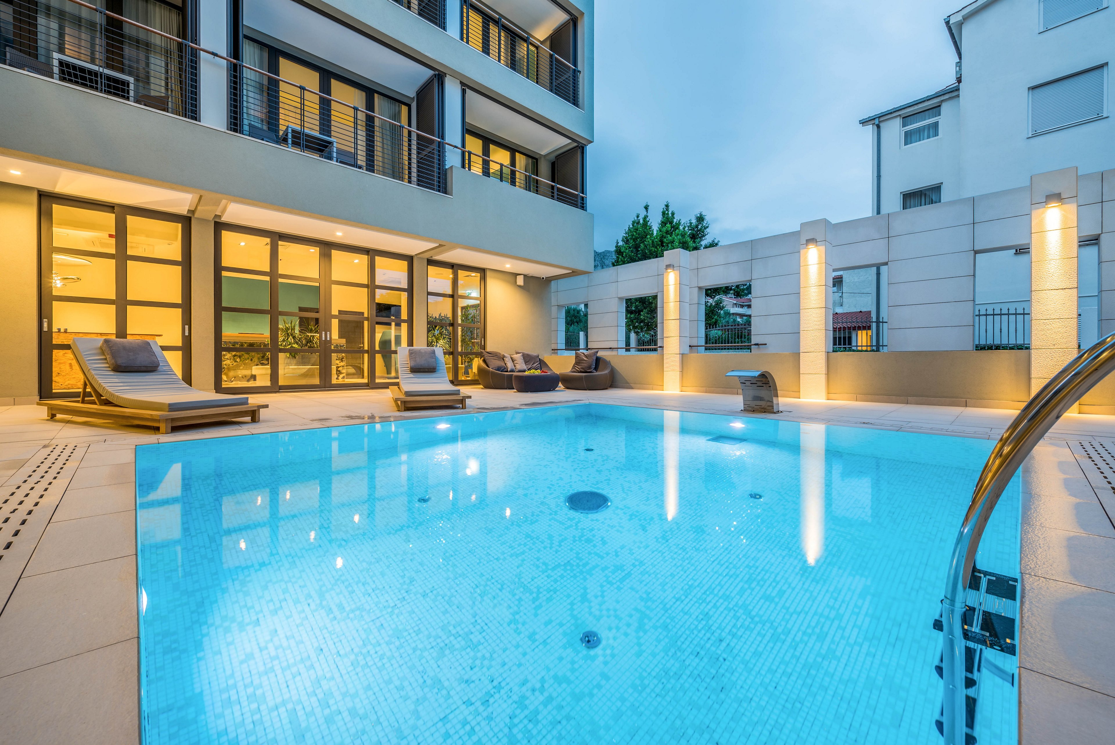A contemporary apartment complex features a glowing outdoor swimming pool at dusk. The pool area is surrounded by lounge chairs and modern architecture, with large windows revealing warmly lit interiors. The tranquil blue water reflects the building’s lights, creating a serene and inviting atmosphere. No visible text or numbers are present in the image.