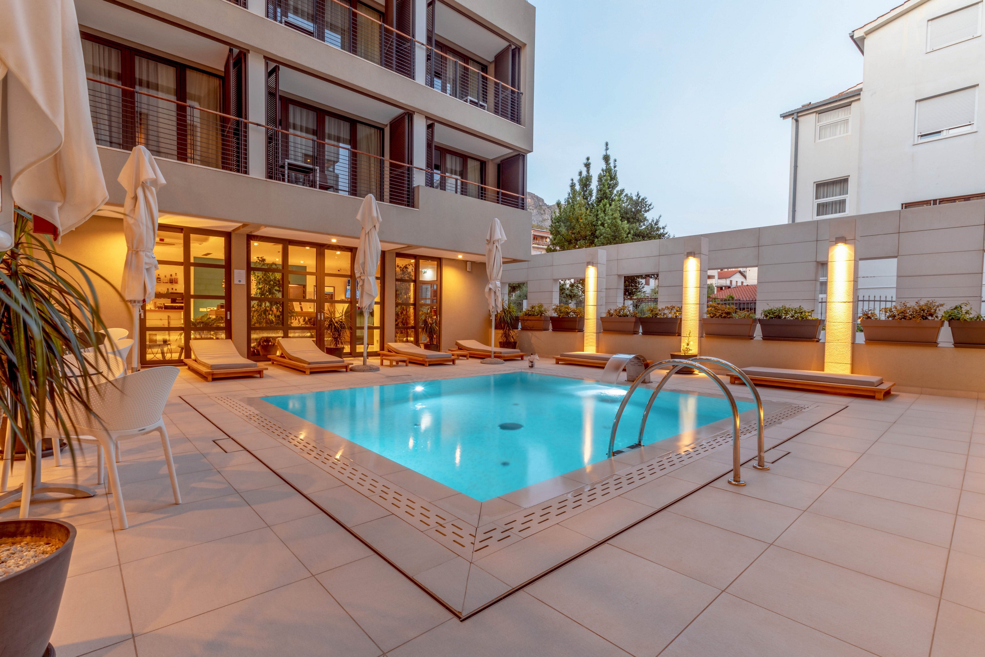 A contemporary outdoor hotel courtyard features a rectangular swimming pool surrounded by lounge chairs and umbrellas. The setting is tranquil, with soft lighting from vertical fixtures and reflections on the water. The area is bordered by modern buildings and potted plants, creating a relaxing urban oasis. No visible people or text are present in the image.
