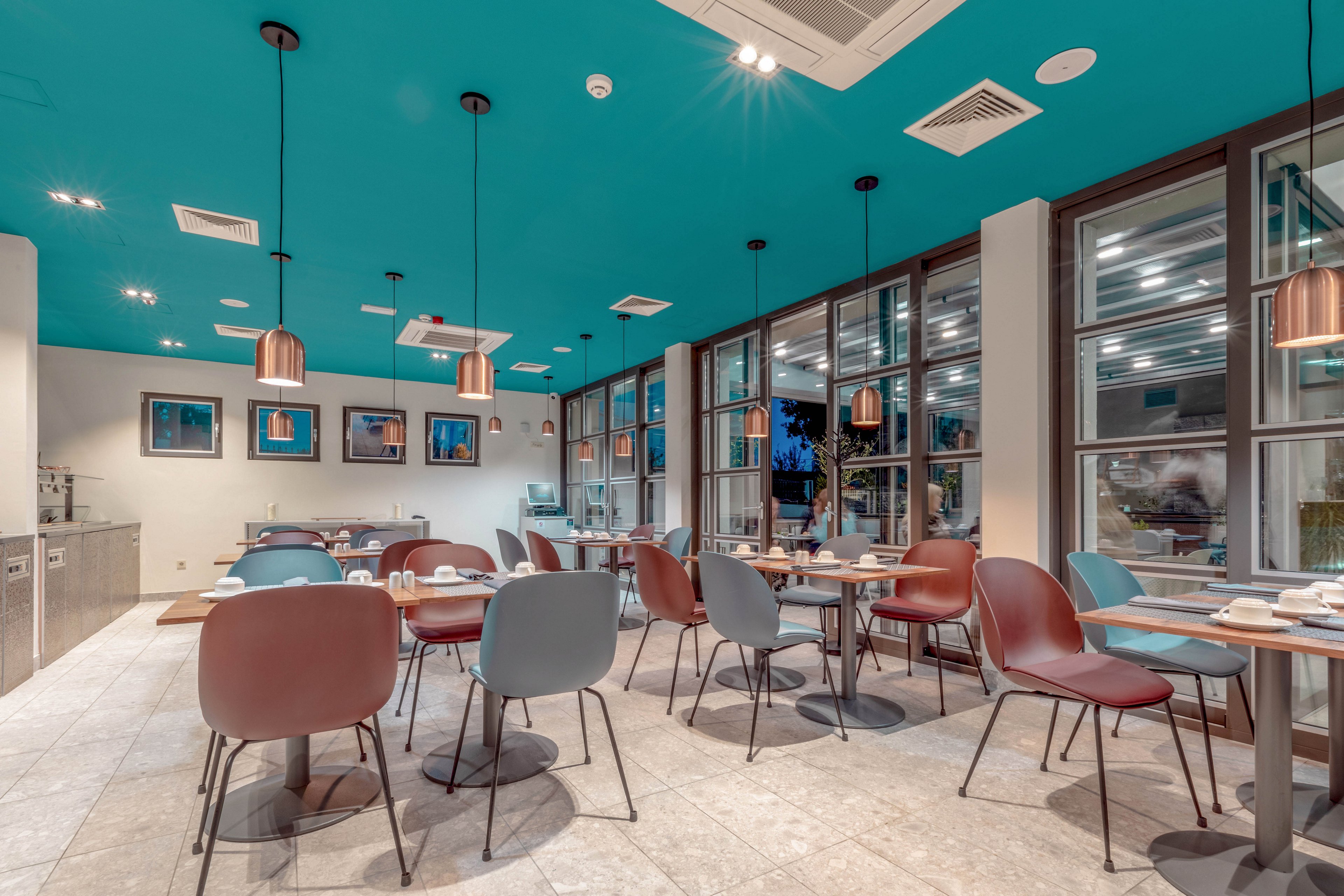 A contemporary restaurant dining area features a striking teal ceiling and copper pendant lights. The space is furnished with minimalist tables and chairs in muted tones, set for service but currently empty. Large windows allow natural light to fill the room, and framed artwork decorates the walls. The overall atmosphere is clean, modern, and inviting.