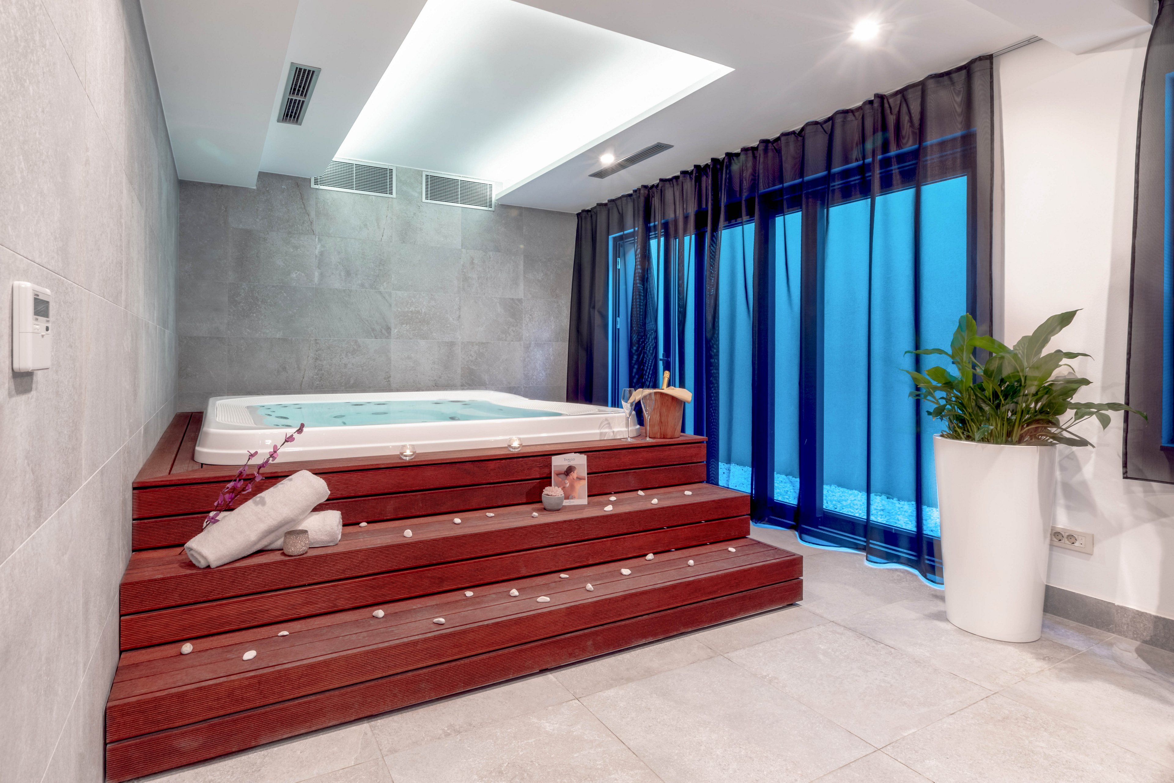 A contemporary indoor spa features a built-in jacuzzi set on wooden steps, surrounded by soft lighting and blue-tinted curtains. Rolled towels, candles, and a wooden bucket add to the relaxing ambiance. The space is accented by a large potted plant and scattered flower petals, creating a tranquil and inviting atmosphere. The overall palette is neutral with pops of blue and green.