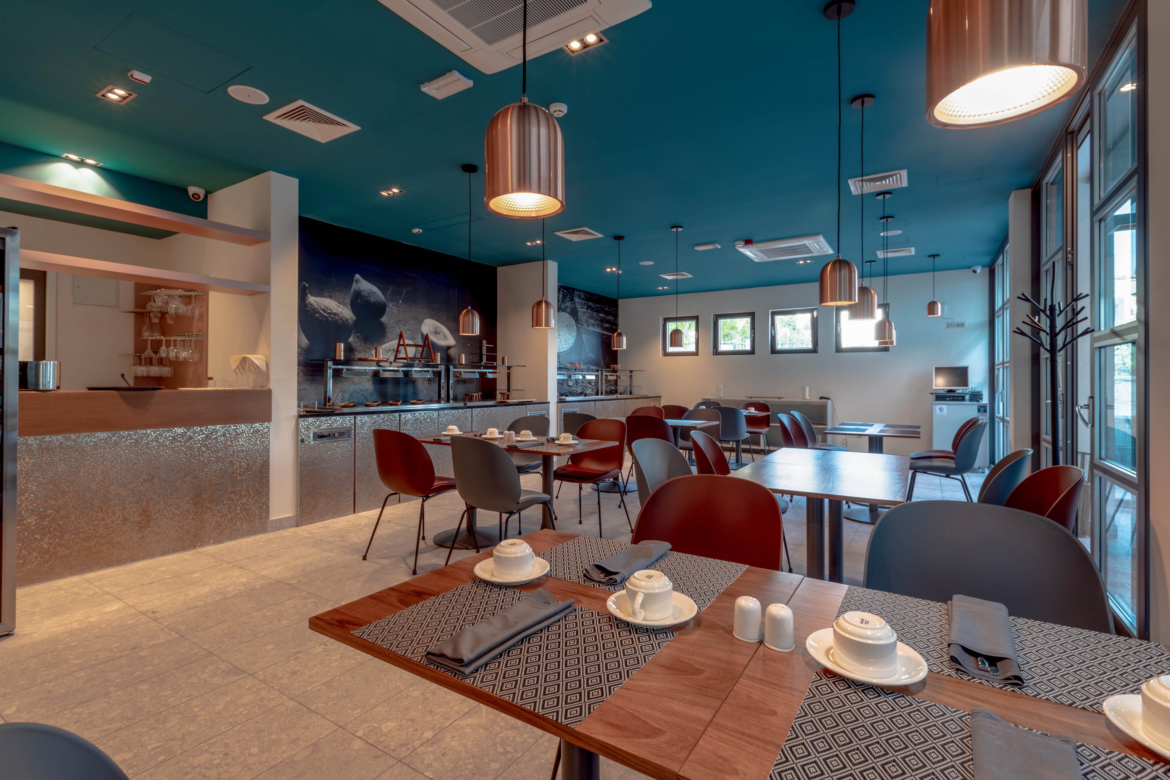 A contemporary restaurant dining area features neatly arranged tables with cups, saucers, and napkins. The setting includes stylish pendant lighting, a teal ceiling, and large windows allowing natural light. The space is empty of people, highlighting the clean and inviting atmosphere. Wall art and metallic accents add to the modern aesthetic.