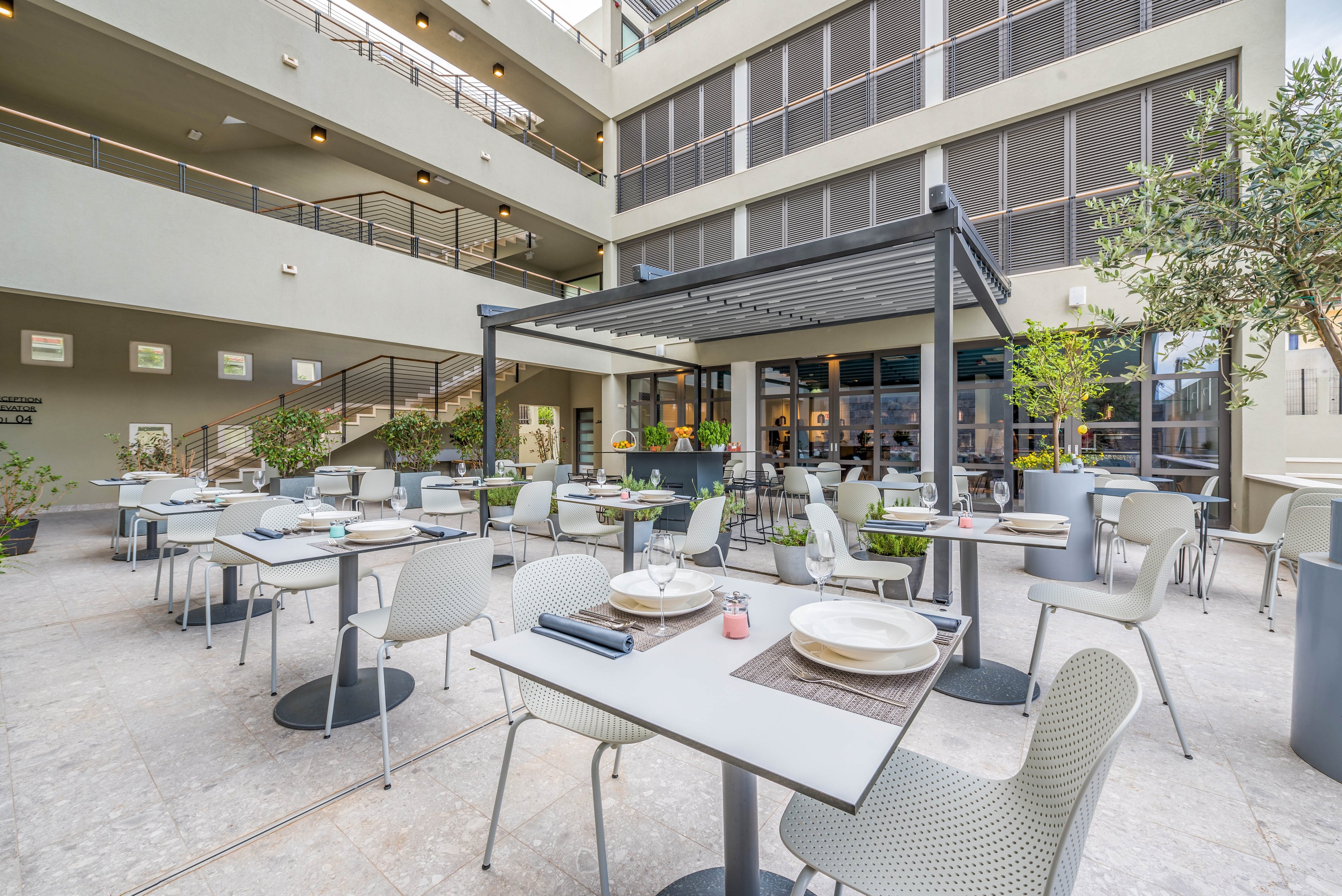 A contemporary outdoor dining area features neatly arranged tables and chairs under a sleek pergola. The setting is part of a multi-story building with large windows and greenery, creating a relaxed and inviting atmosphere. Table settings include plates, glasses, and napkins, with no people present. The color palette is neutral with touches of green from plants.