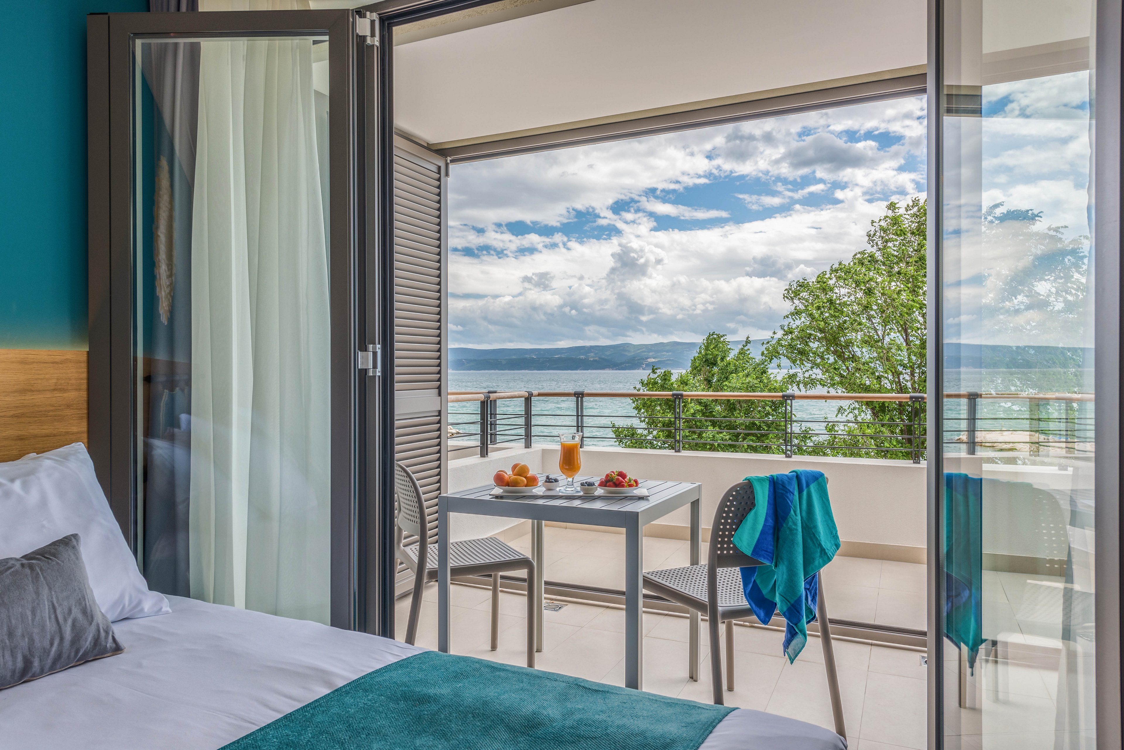 A contemporary hotel room opens onto a private balcony overlooking the sea. The balcony features a small table set with drinks and fruit, and two chairs, one draped with a blue towel. The scene is bright and inviting, with turquoise accents and a tranquil atmosphere. No visible text or numbers are present in the image.