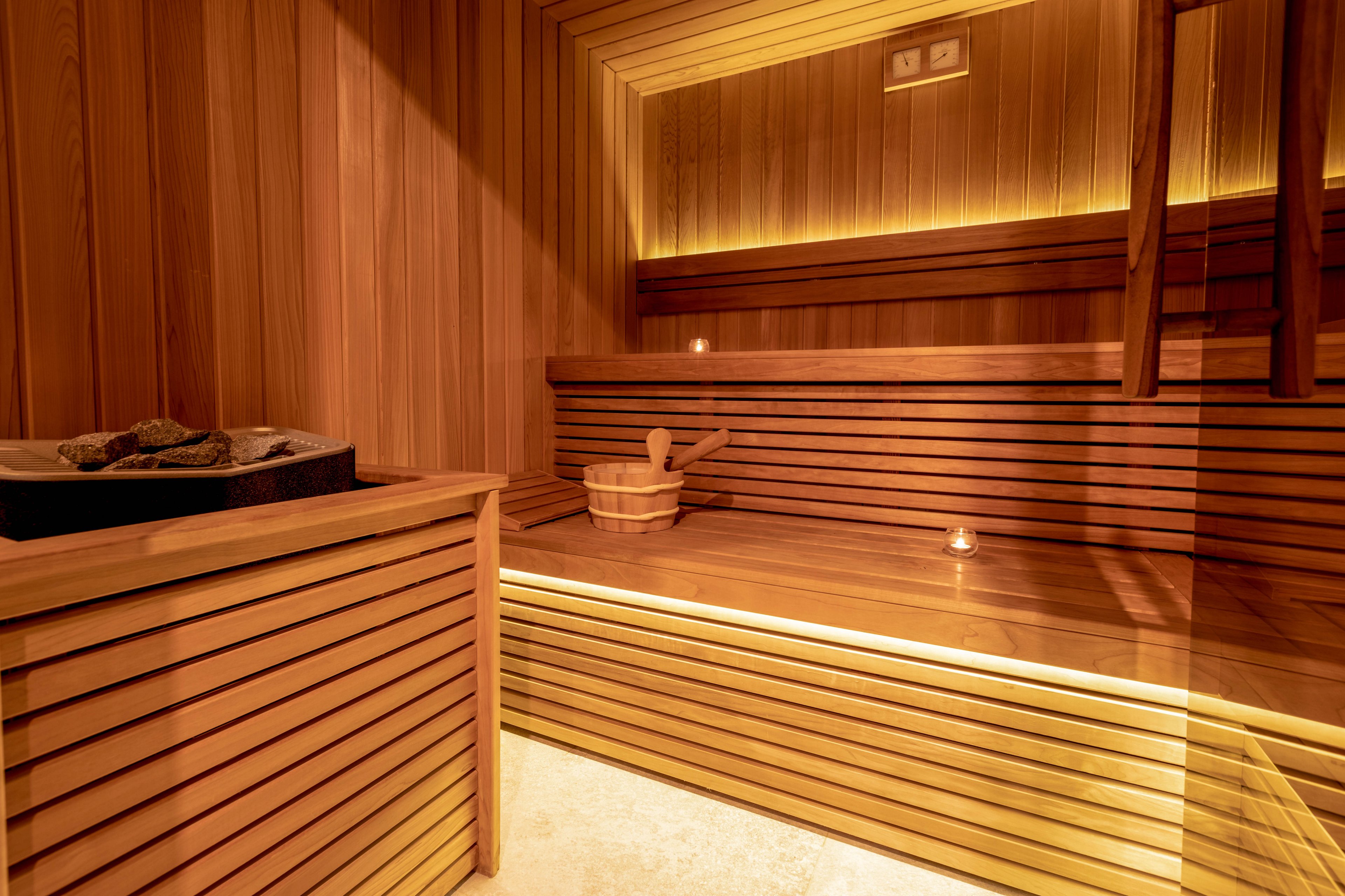 A contemporary sauna room features smooth wooden paneling and soft, ambient lighting. A wooden bucket and ladle are placed on the bench, with a candle adding to the tranquil atmosphere. The sauna heater with stones is visible in the corner, enhancing the authentic spa experience. The setting is empty, emphasizing relaxation and calm.