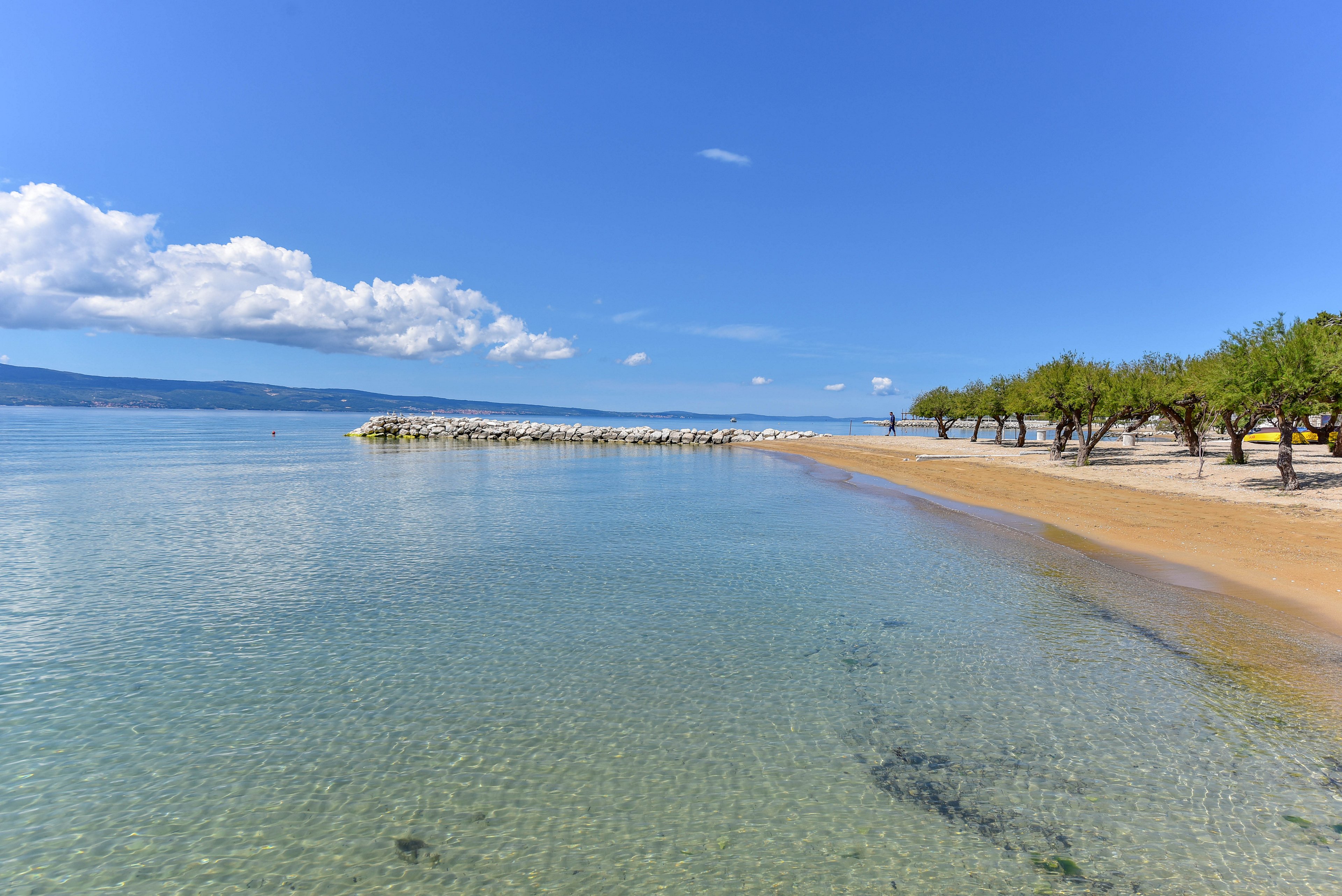 A tranquil sandy beach stretches along a calm, clear shoreline under a bright blue sky. The water is transparent, revealing the sandy bottom, while a line of small trees provides shade in the distance. A stone breakwater extends into the water, and a few clouds dot the sky, enhancing the peaceful atmosphere. No people are visible, emphasizing the serene and undisturbed setting.