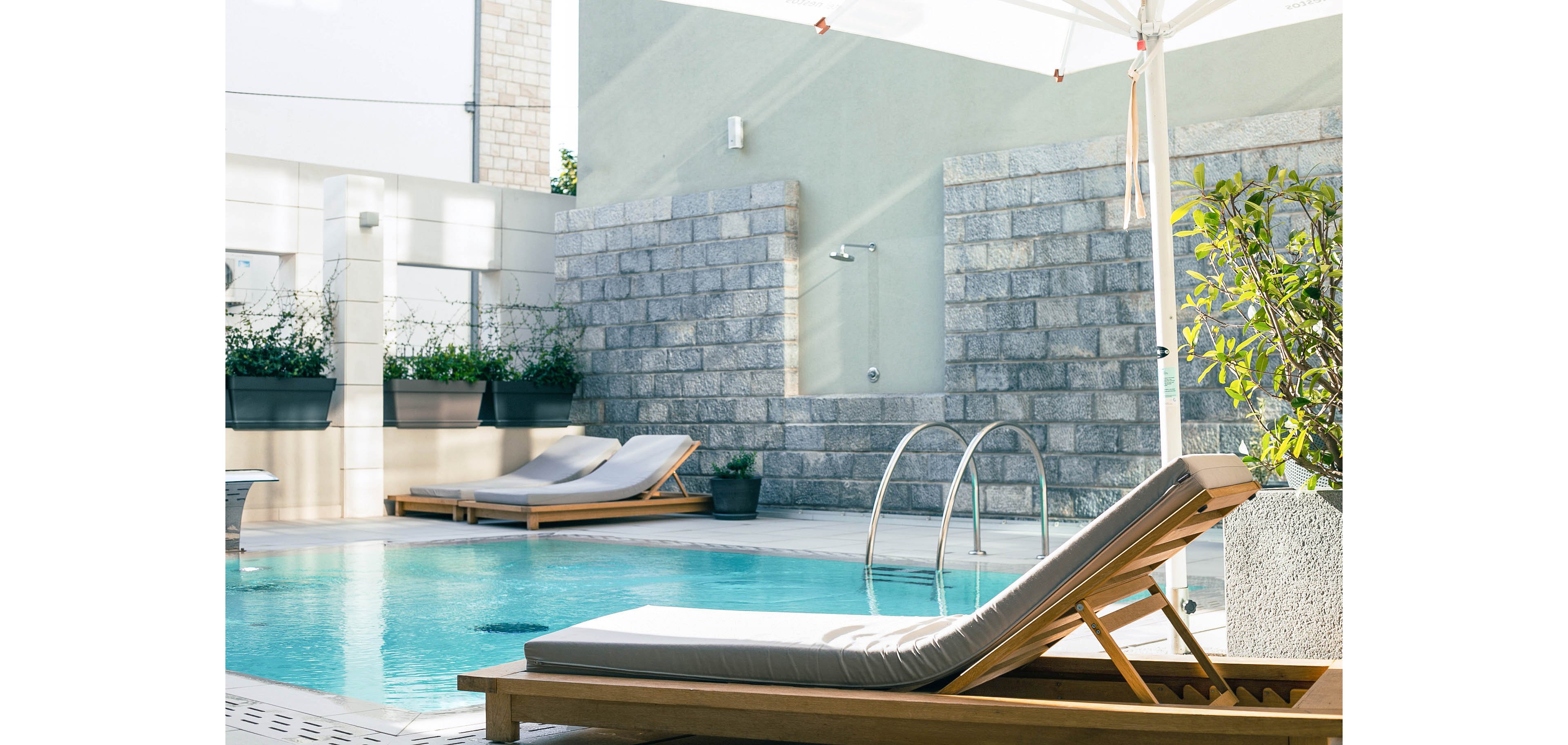 A serene outdoor pool area features two cushioned sun loungers beside a clear blue swimming pool. The setting is modern, with stone walls, potted plants, and a pool ladder visible. Soft natural light and neutral tones create a relaxing, inviting atmosphere. No people are present, emphasizing tranquility and privacy.