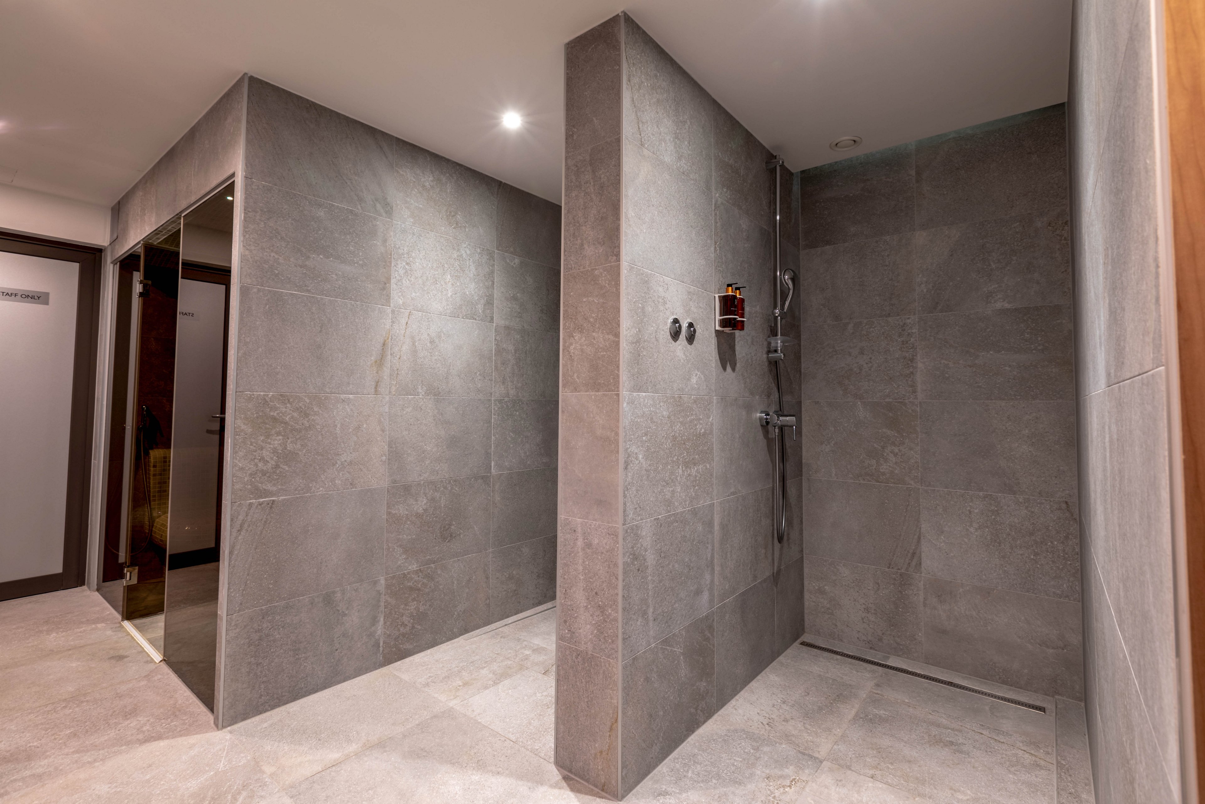 A spacious, contemporary communal shower area features large gray tiles covering the walls and floor. The setting is clean and minimalistic, with a walk-in shower and a glass door visible in the background. Overhead lighting creates a bright and inviting atmosphere, emphasizing the sleek, modern design. No visible text or numbers are present in the image.