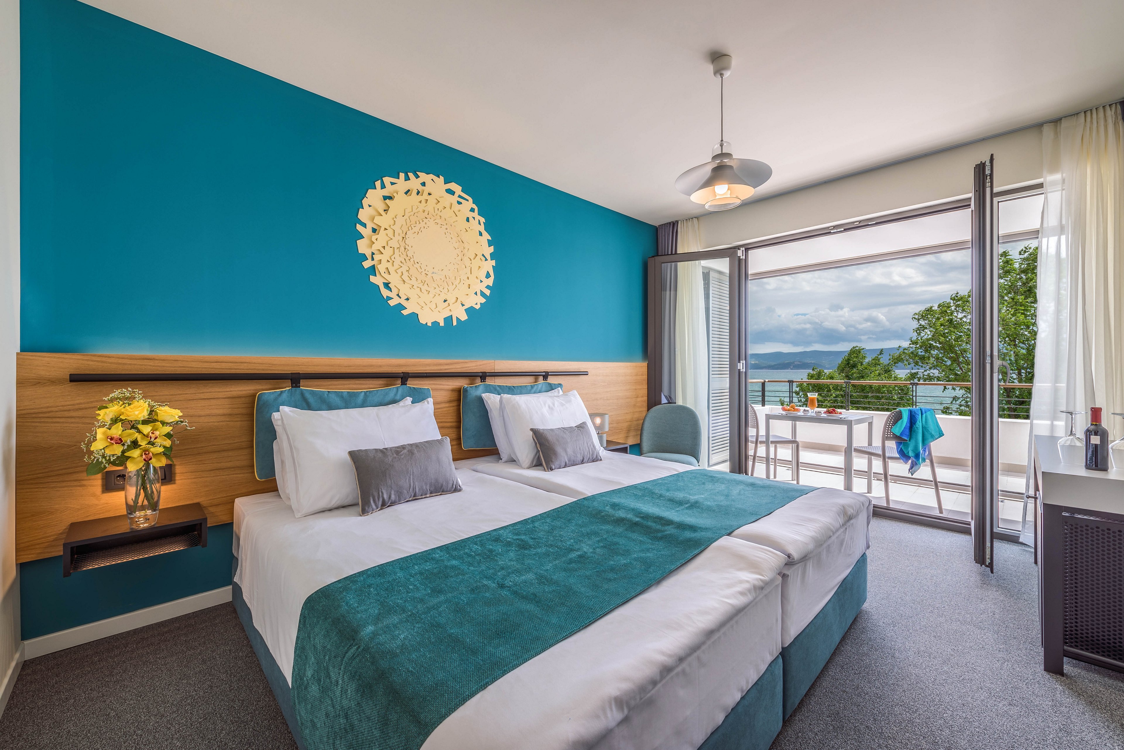 A contemporary hotel room features two twin beds with teal accents and a decorative wall piece above the headboard. The space is bright and airy, with a large glass door opening to a balcony overlooking the sea and lush greenery. Fresh flowers and a bottle of wine add a welcoming touch, while the room's clean lines and teal color palette create a relaxing atmosphere. No visible text or numbers are present in the image.