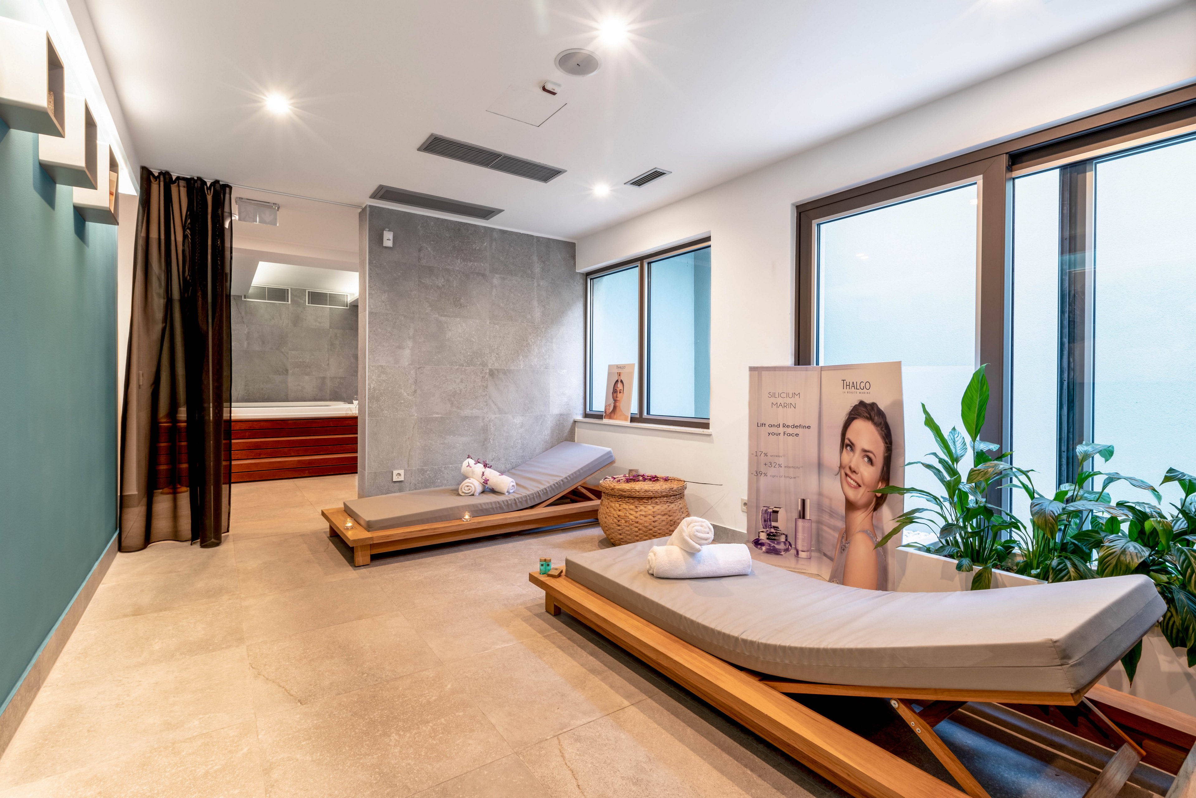 A contemporary spa relaxation room features two cushioned lounge chairs, a basket with towels, and a large potted plant near expansive windows. The setting is bright and tranquil, with neutral tones and soft lighting enhancing the serene atmosphere. A visible product display shows a partially obscured image of a woman and the text 'SABO,' adding a subtle commercial element. The space includes a glimpse of a hot tub area in the background, contributing to the overall wellness ambiance.