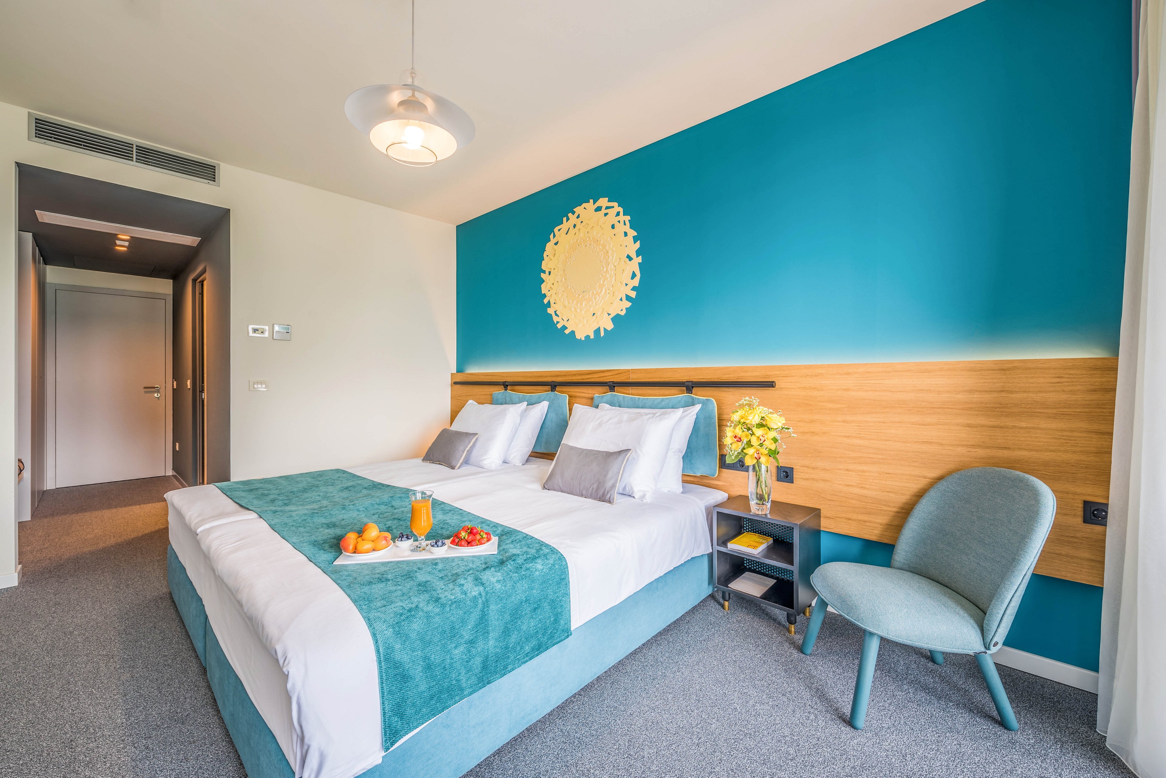 A contemporary hotel room features a double bed with teal and white bedding, complemented by a teal accent wall and wooden headboard. The setting includes a bedside table with a vase of yellow flowers, a chair, and a tray with orange juice and fruit on the bed. The room is brightly lit with natural light and has a clean, inviting atmosphere. No visible text or numbers are present in the image.