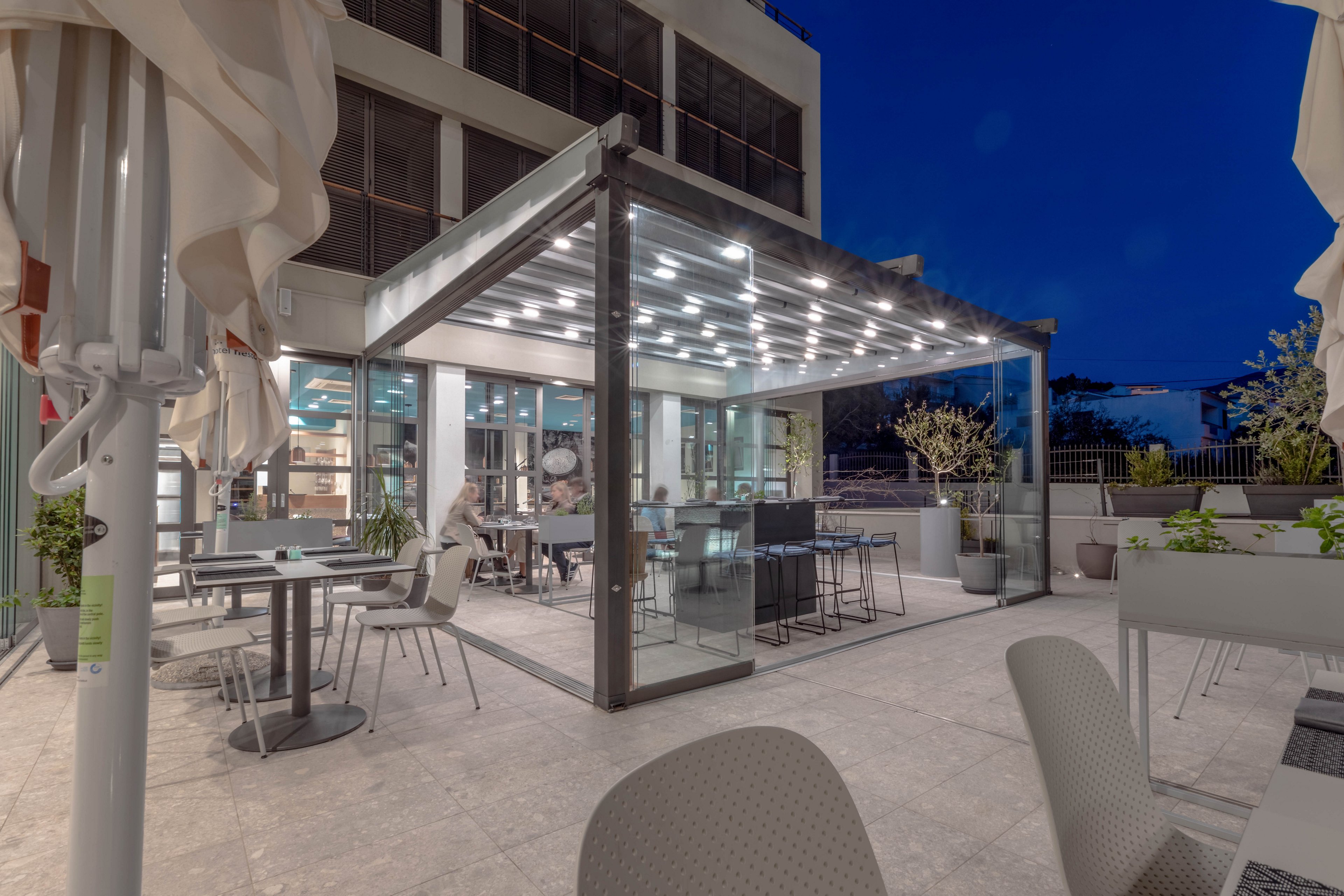 The image shows a contemporary outdoor restaurant patio with tables and chairs arranged under a glass canopy. The setting is evening, with artificial lighting illuminating the area and a clear blue sky in the background. A few people are seated, enjoying a relaxed atmosphere. The visuals highlight sleek furniture, potted plants, and a clean, minimalist design. No visible text or numeric values are present.