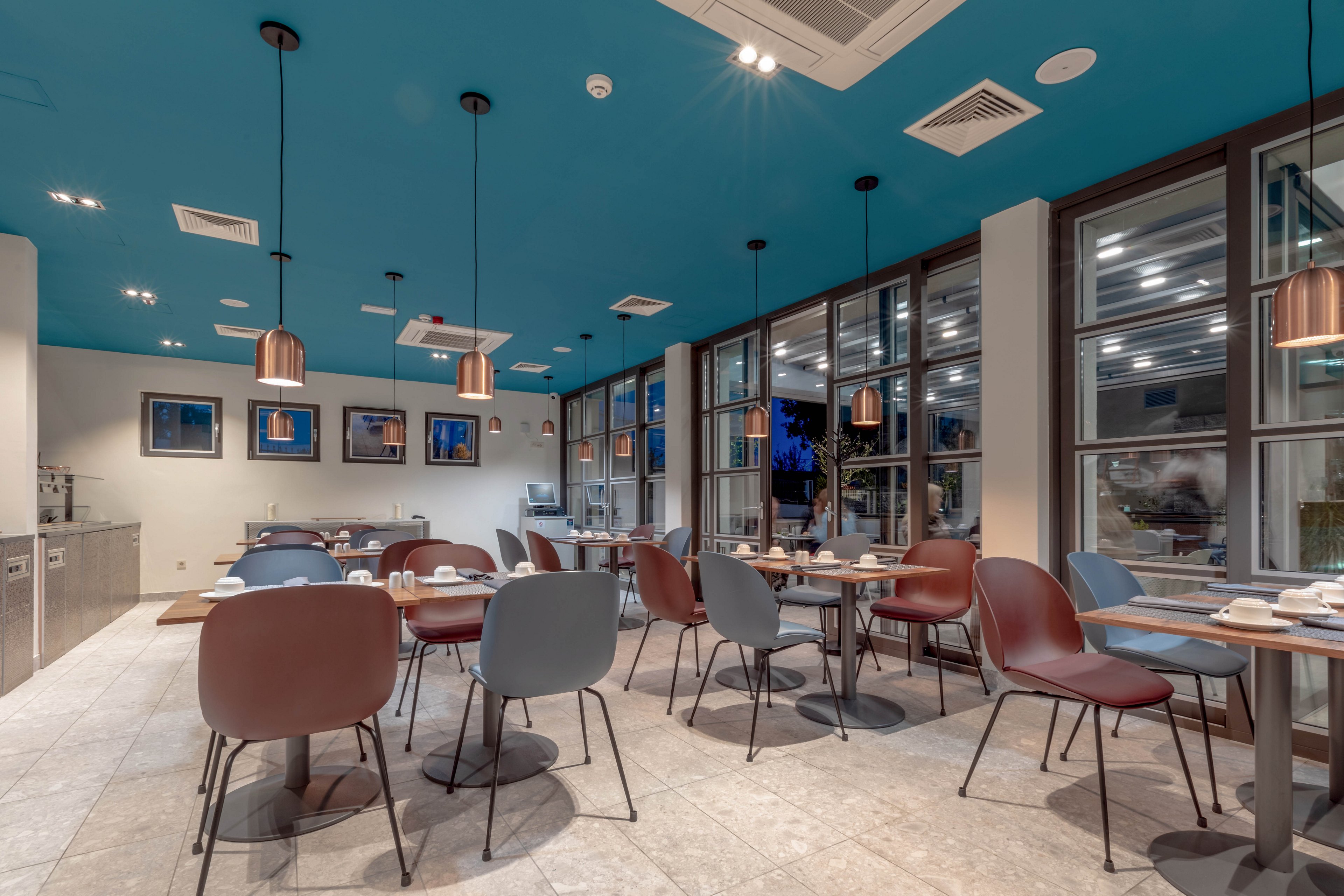 A contemporary dining space features a striking blue ceiling and copper pendant lights. The room is furnished with minimalist chairs and tables, set for breakfast or casual dining. Large windows allow natural light to fill the area, while framed artwork decorates the walls. The setting is clean, inviting, and designed for comfort.