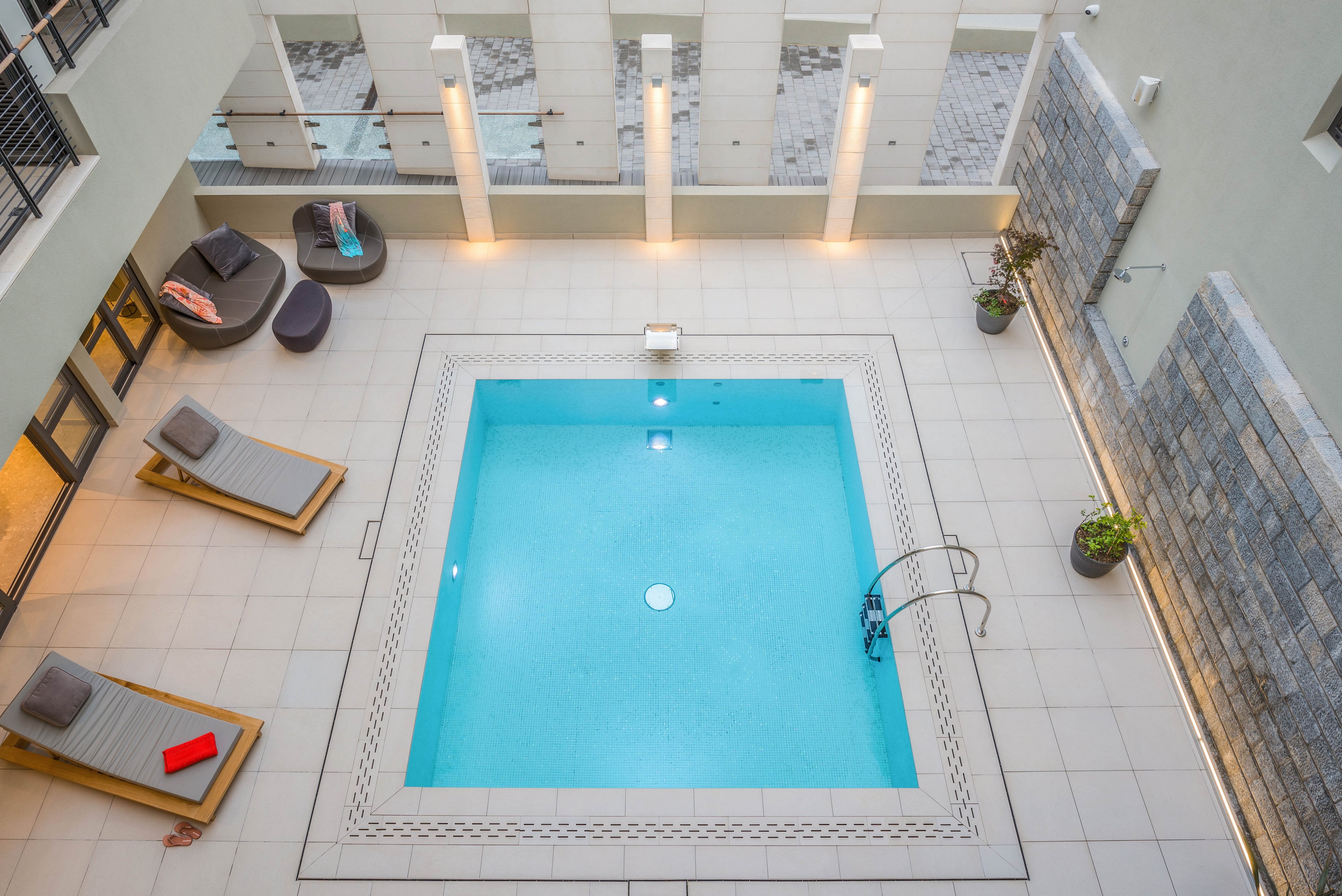 A square indoor swimming pool is centered in a clean, contemporary space with light tile flooring. Surrounding the pool are cushioned lounge chairs, a small seating area, and potted plants, creating a relaxing atmosphere. The setting is well-lit with wall lights and features a minimalist, modern design with neutral tones and a pop of color from a red book.