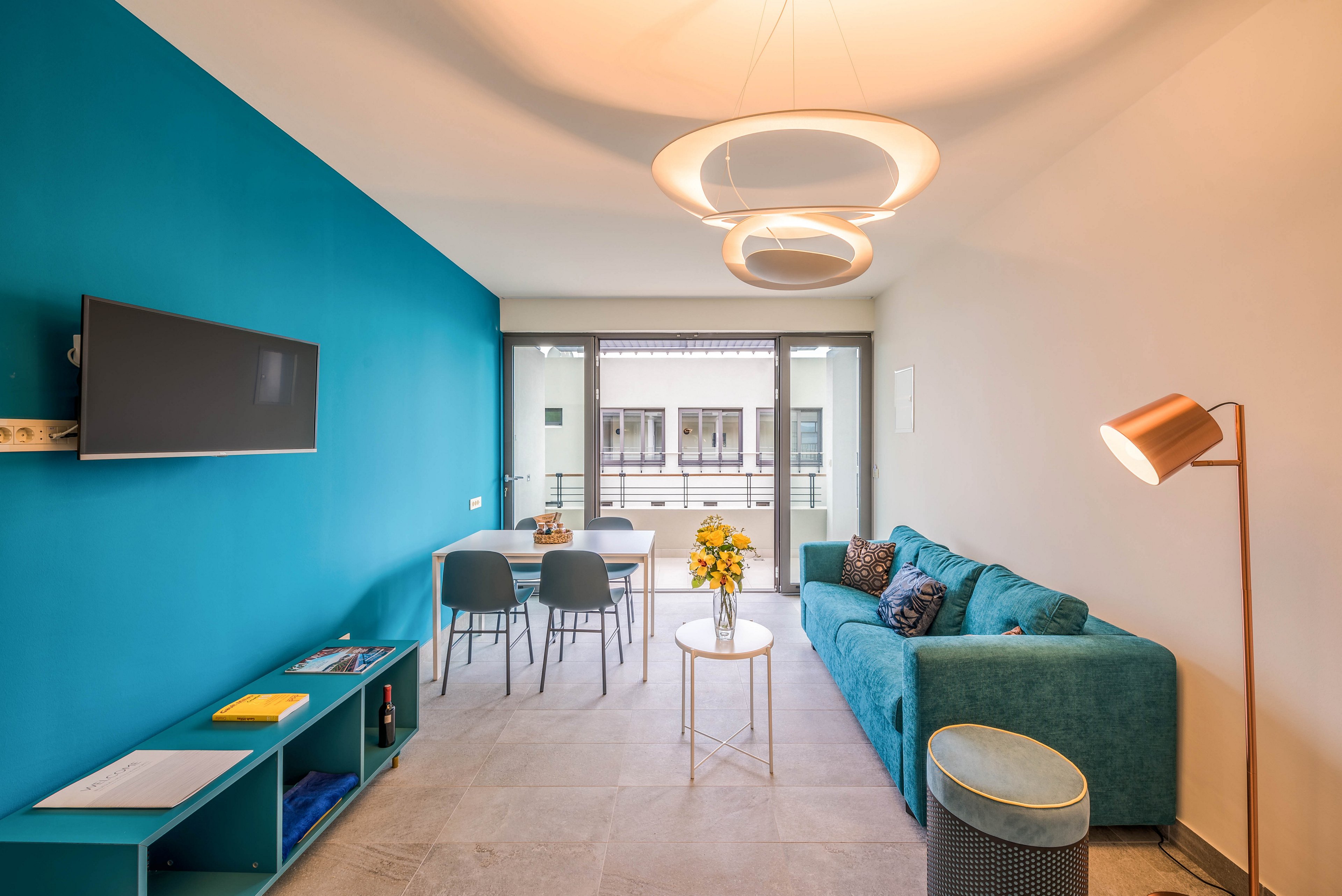 A contemporary living room features a teal accent wall, matching sofa, and a mounted flat-screen television. The space includes a dining table with four chairs near a large window leading to a balcony, allowing natural light to fill the room. Decorative lighting fixtures and a floor lamp add a stylish touch, while a vase of yellow flowers brightens the coffee table. The overall palette is teal, white, and neutral tones, creating a fresh and inviting atmosphere.