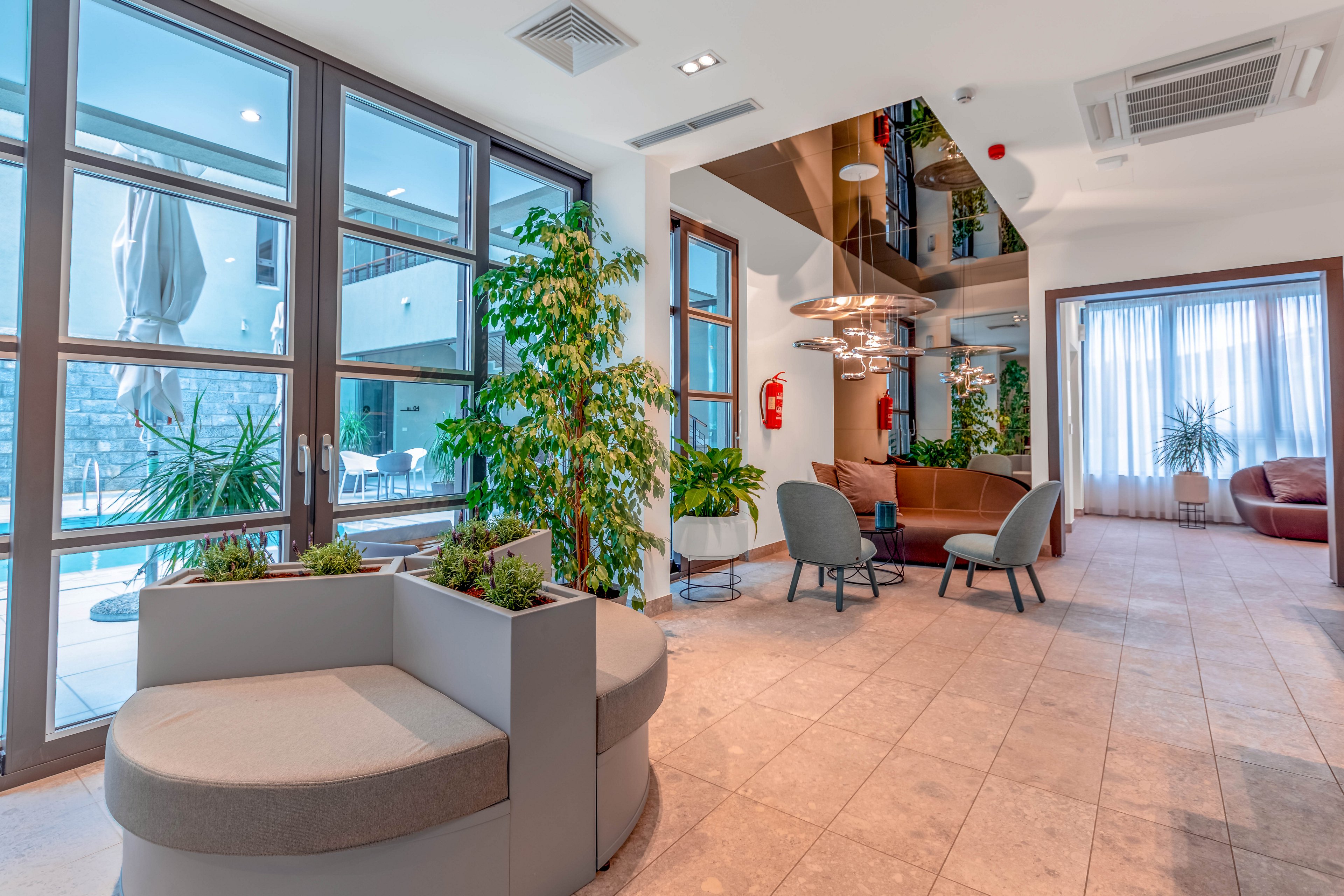 A contemporary hotel lounge features stylish seating arrangements and large floor-to-ceiling windows overlooking an outdoor pool area. The space is decorated with indoor plants, neutral-toned furniture, and pendant lighting, creating a welcoming and airy atmosphere. Natural light fills the room, highlighting the clean lines and modern design elements. No visible text or numbers are present in the image.