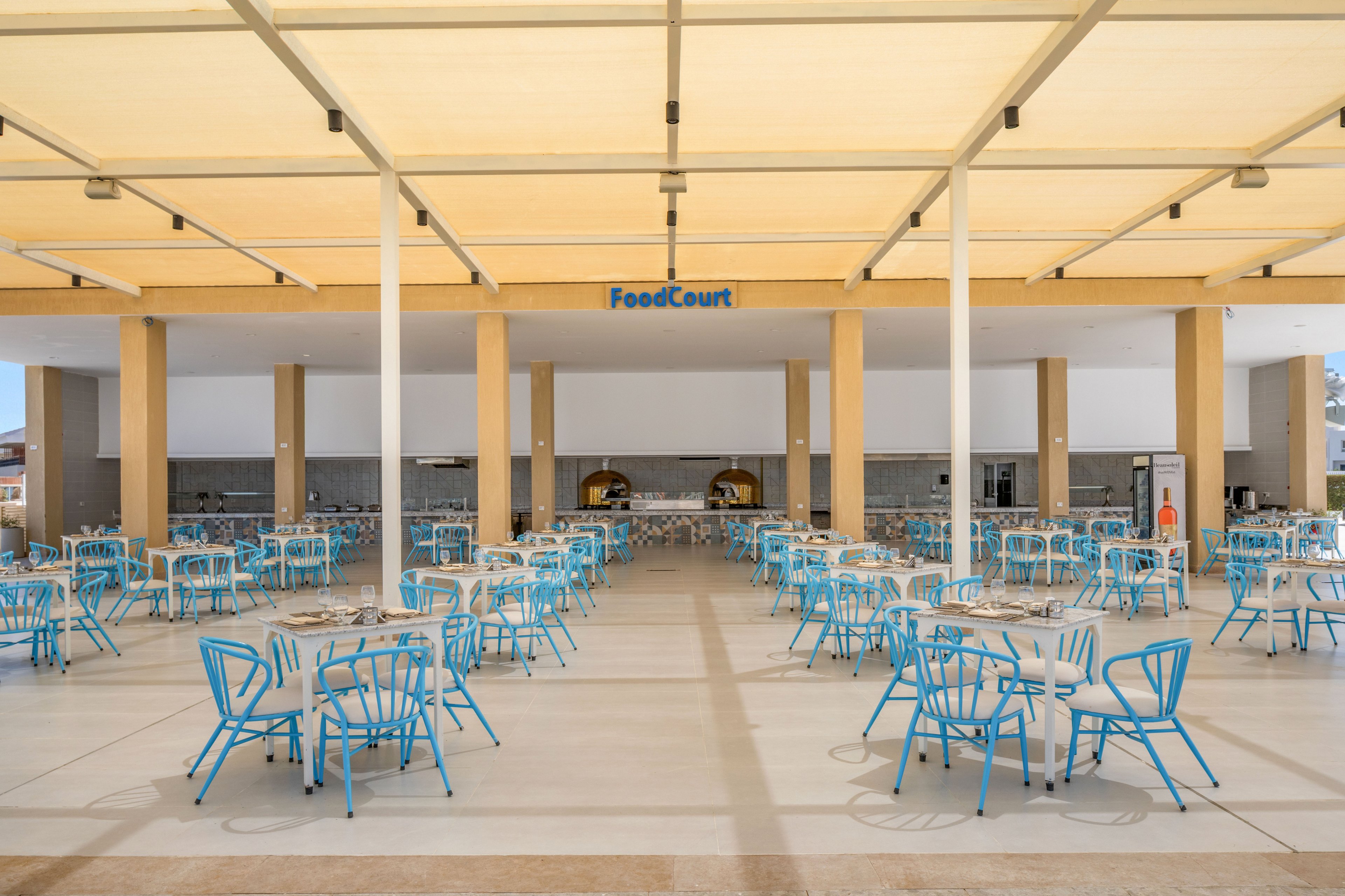 An open air food court features rows of empty tables and bright blue chairs under a large beige canopy. The space is clean and orderly, with a modern minimalist design and neutral tones contrasted by the colorful seating. In the background, food service counters and ovens are visible, suggesting a casual dining environment. The word "FoodCourt" is clearly displayed above the central serving area.