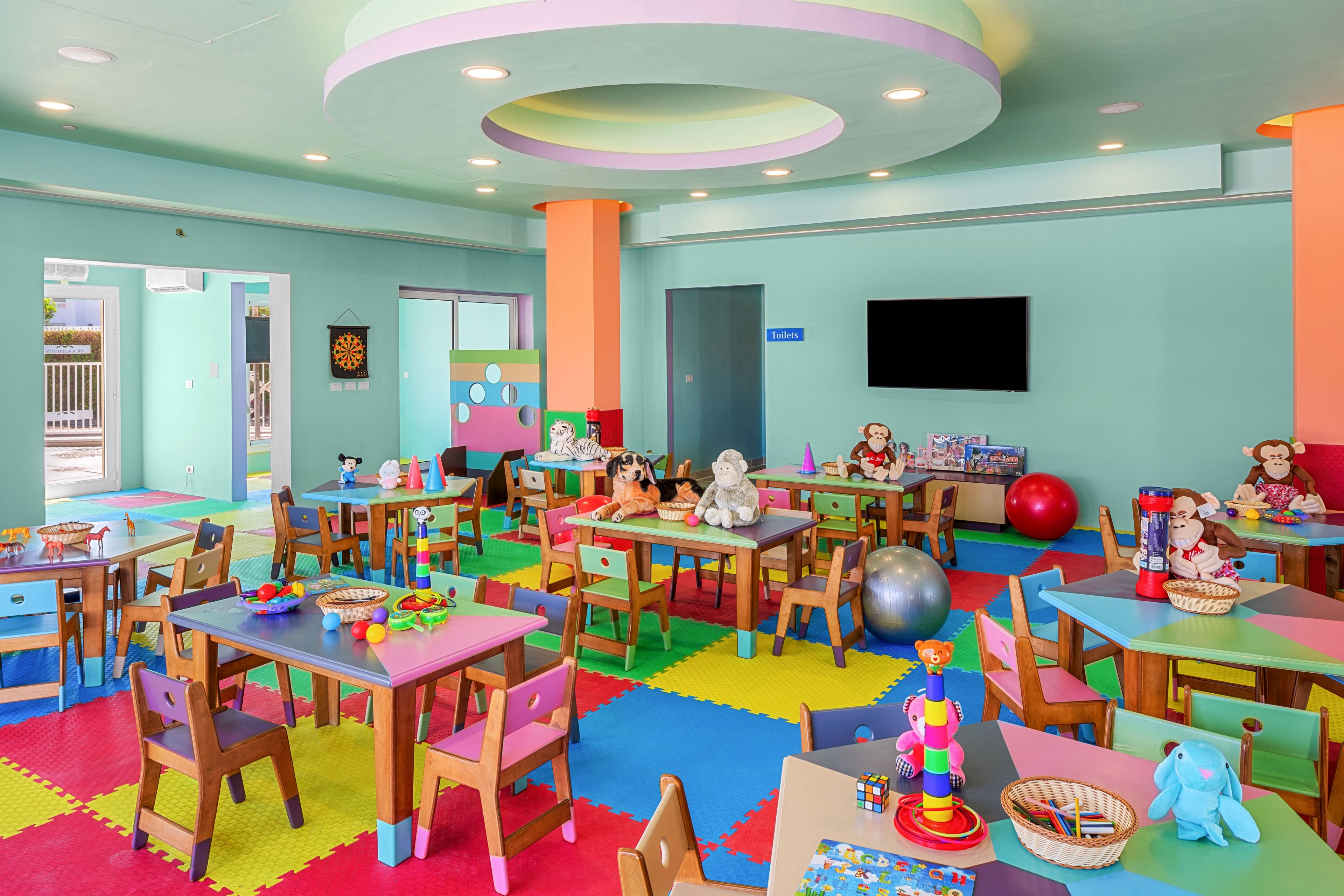 A bright, colorful playroom is filled with small tables and chairs arranged neatly around the room. The space features foam floor tiles in vibrant colors, pastel walls, and a circular ceiling detail with recessed lighting. Toys, stuffed animals, puzzles, and art supplies are placed on the tables and shelves, creating a playful and inviting environment. Large windows and open doors let in natural light, while a wall-mounted television and decorative wall panels add to the modern feel of the children's area.