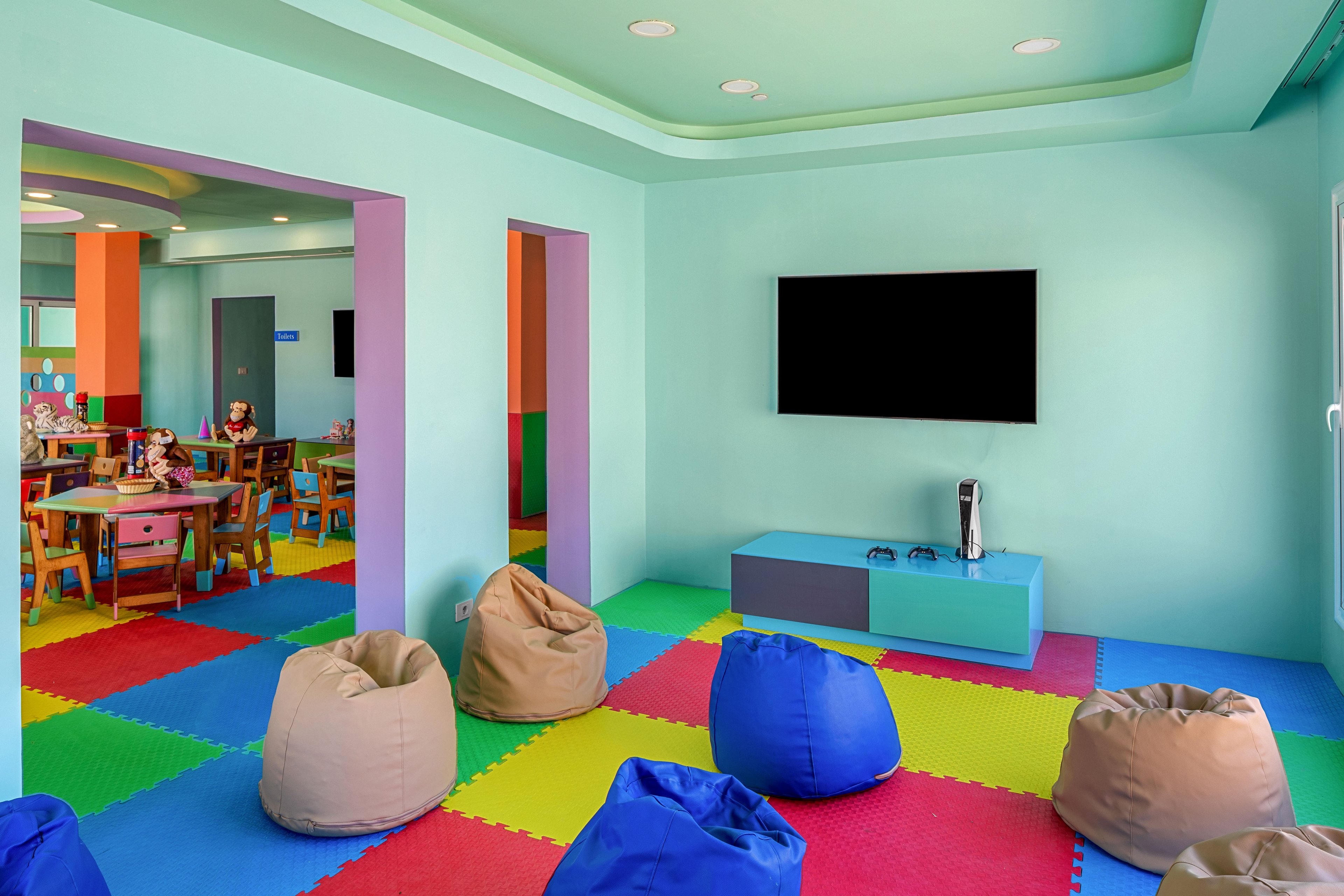 A bright children’s playroom features a large flat-screen television mounted on a pastel wall above a low media console. The floor is covered with interlocking foam tiles in vibrant colors, and several bean bag chairs are arranged in the center of the room. In the background, another play area with small tables, chairs, and toys is visible through open doorways. The overall space feels cheerful and inviting, designed for kids to relax, play, and learn.