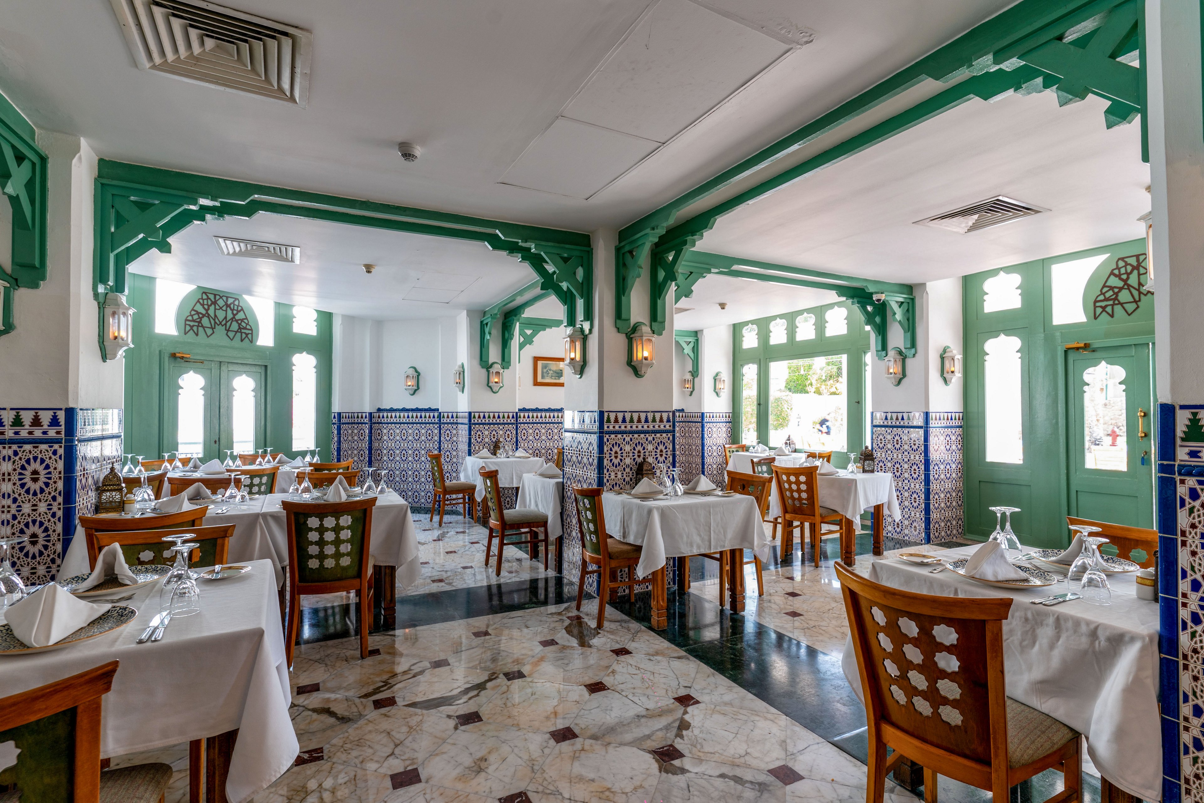 Elegant restaurant dining room with neatly arranged tables and wooden chairs. White tablecloths, folded napkins, and glassware are set for guests in a bright, airy space. Green architectural details frame windows and doors, complemented by patterned blue and white wall tiles. Natural light streams in, highlighting the polished floor and inviting atmosphere.