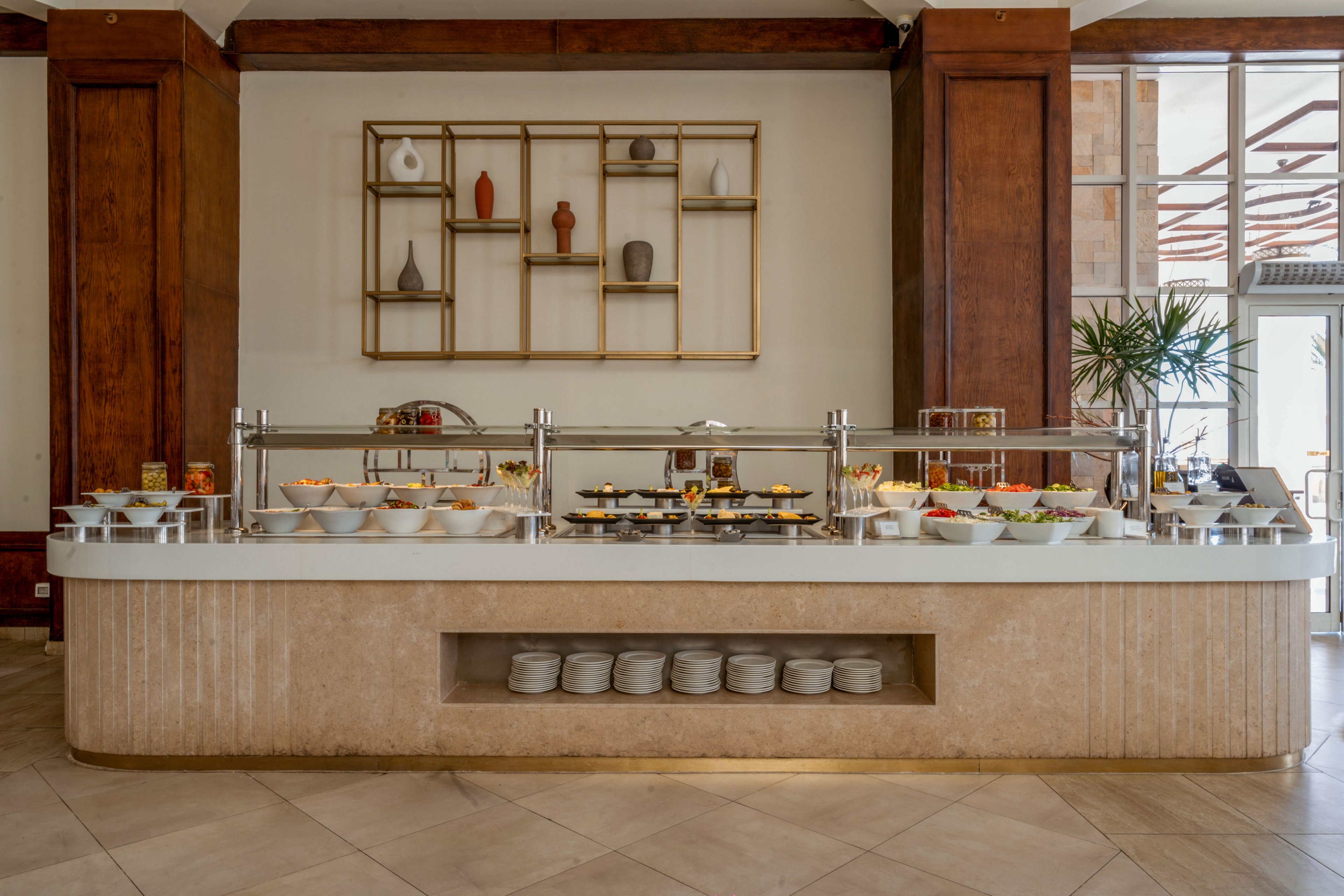 A spacious buffet counter is set up in a bright, modern dining area. Various bowls and platters of food are neatly arranged under a glass sneeze guard, with stacks of plates ready for guests. The background features wooden wall panels and a decorative shelving unit with minimalist vases. Natural light enters from the side, highlighting the clean, neutral tones and contemporary design.