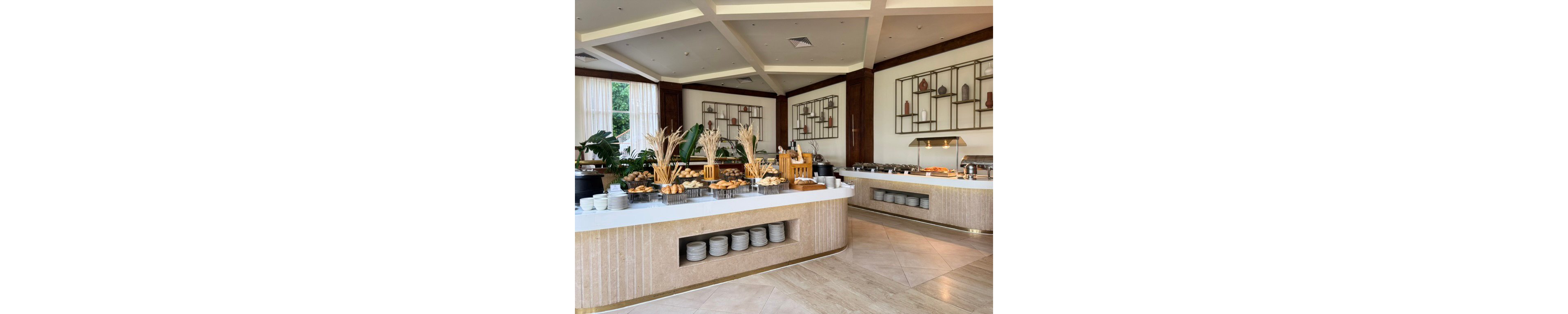 A spacious, modern dining area features a well-organized breakfast buffet with pastries, bread, and hot dishes. White countertops and light stone finishes contrast with dark wooden wall panels and geometric shelving displaying decorative vases. Large windows with sheer curtains let in natural light, highlighting plants and neatly stacked plates beneath the counters. The overall atmosphere is clean, inviting, and designed for a comfortable hotel dining experience.
