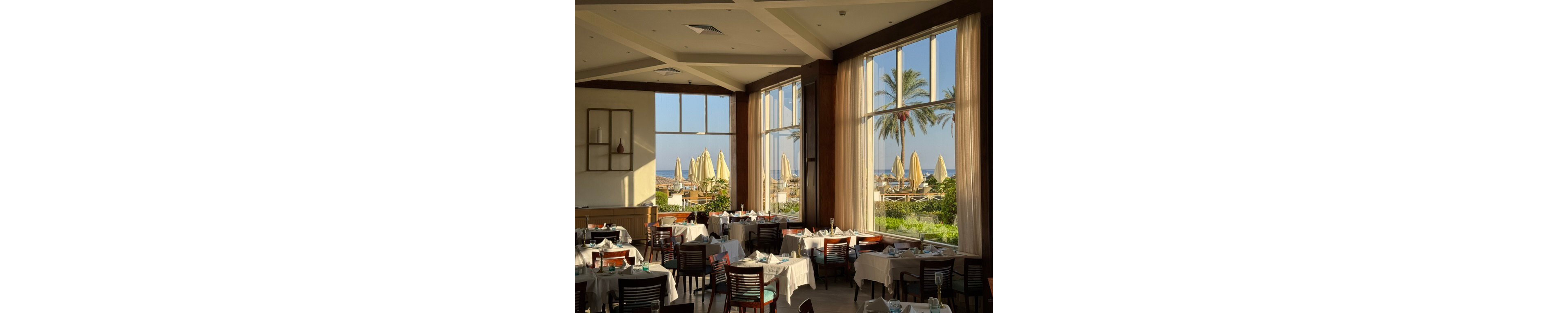 Bright natural light fills a spacious restaurant with neatly arranged tables and wooden chairs. Large floor-to-ceiling windows reveal an outdoor terrace with sun loungers, umbrellas, palm trees, and a distant view of the sea. White tablecloths and simple decor create an elegant yet relaxed dining atmosphere. The scene suggests a coastal resort setting during a sunny day.