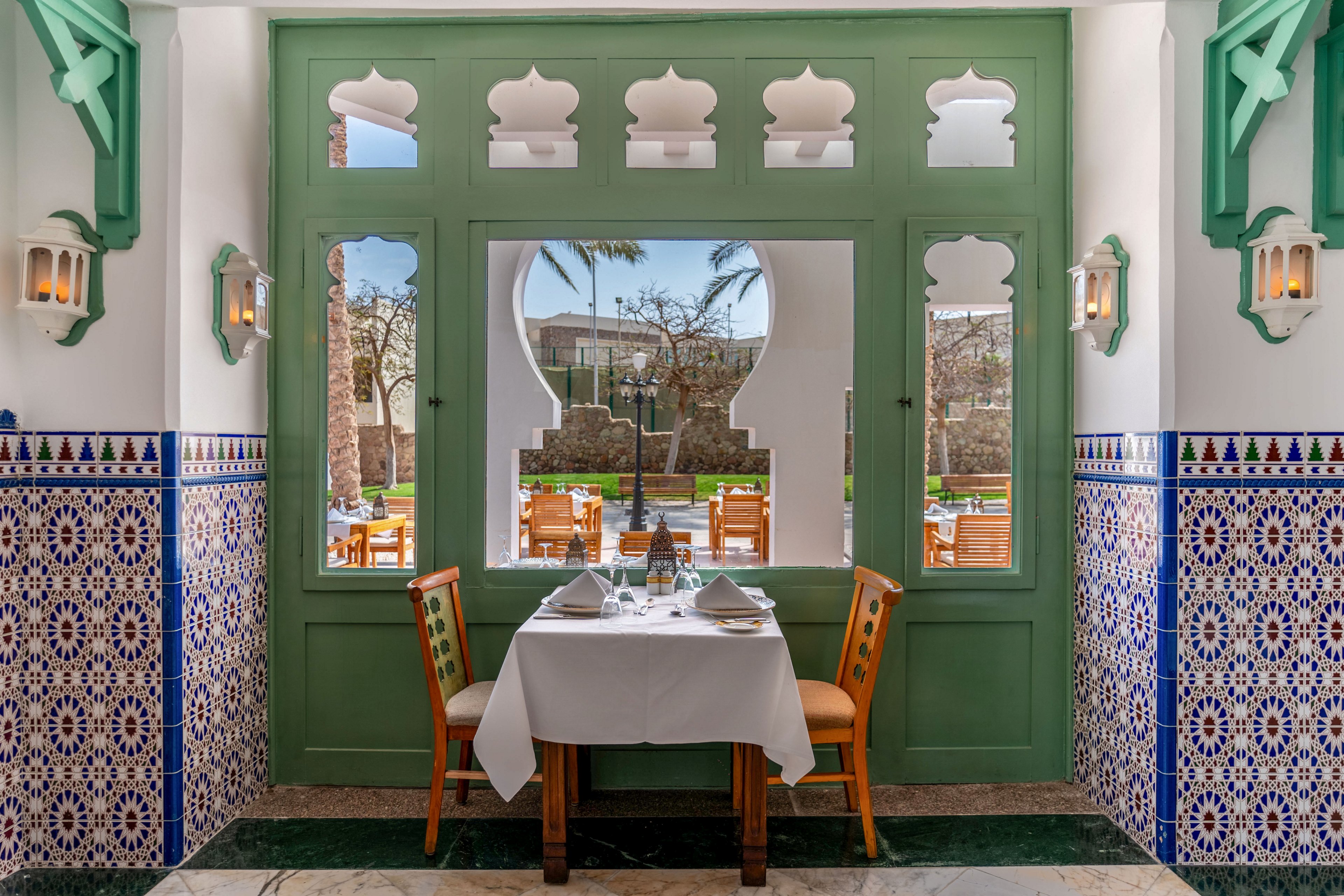 A neatly set dining table for two stands inside a restaurant, framed by ornate green window panels. Colorful geometric mosaic tiles cover the lower walls, adding intricate detail to the space. Through the arched window openings, wooden chairs and tables are visible on a sunny terrace with palm trees and garden views. The scene suggests a relaxed, upscale dining experience in a warm, inviting setting.