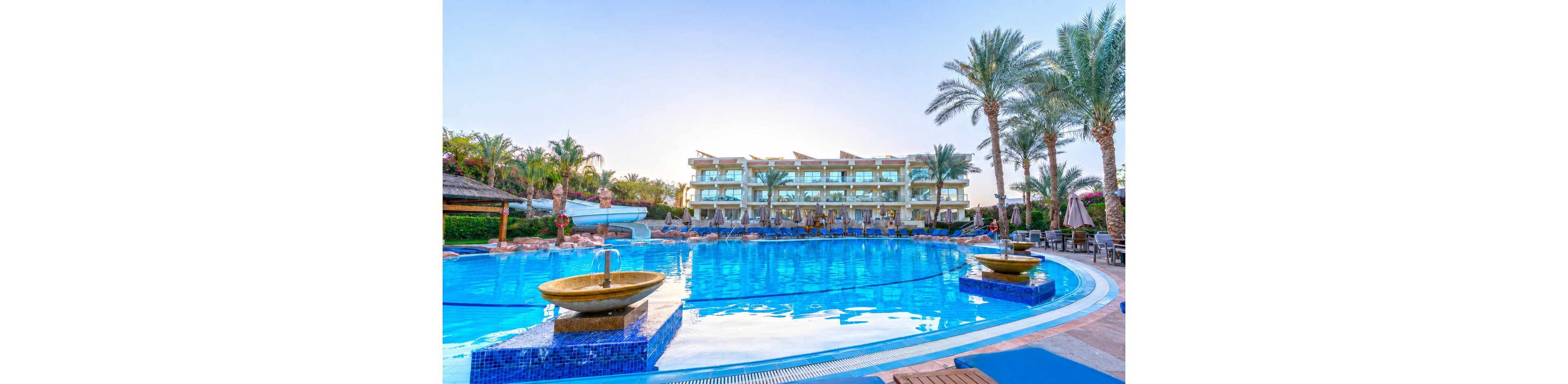 A large, curved swimming pool dominates the foreground of a luxury resort scene at sunset. In the background, a multi-story hotel building with balconies overlooks the pool area, surrounded by lush palm trees and tropical landscaping. Decorative water features and sun loungers line the poolside, creating a relaxing vacation atmosphere. The clear blue water reflects the pastel evening sky, emphasizing a tranquil and inviting holiday setting.