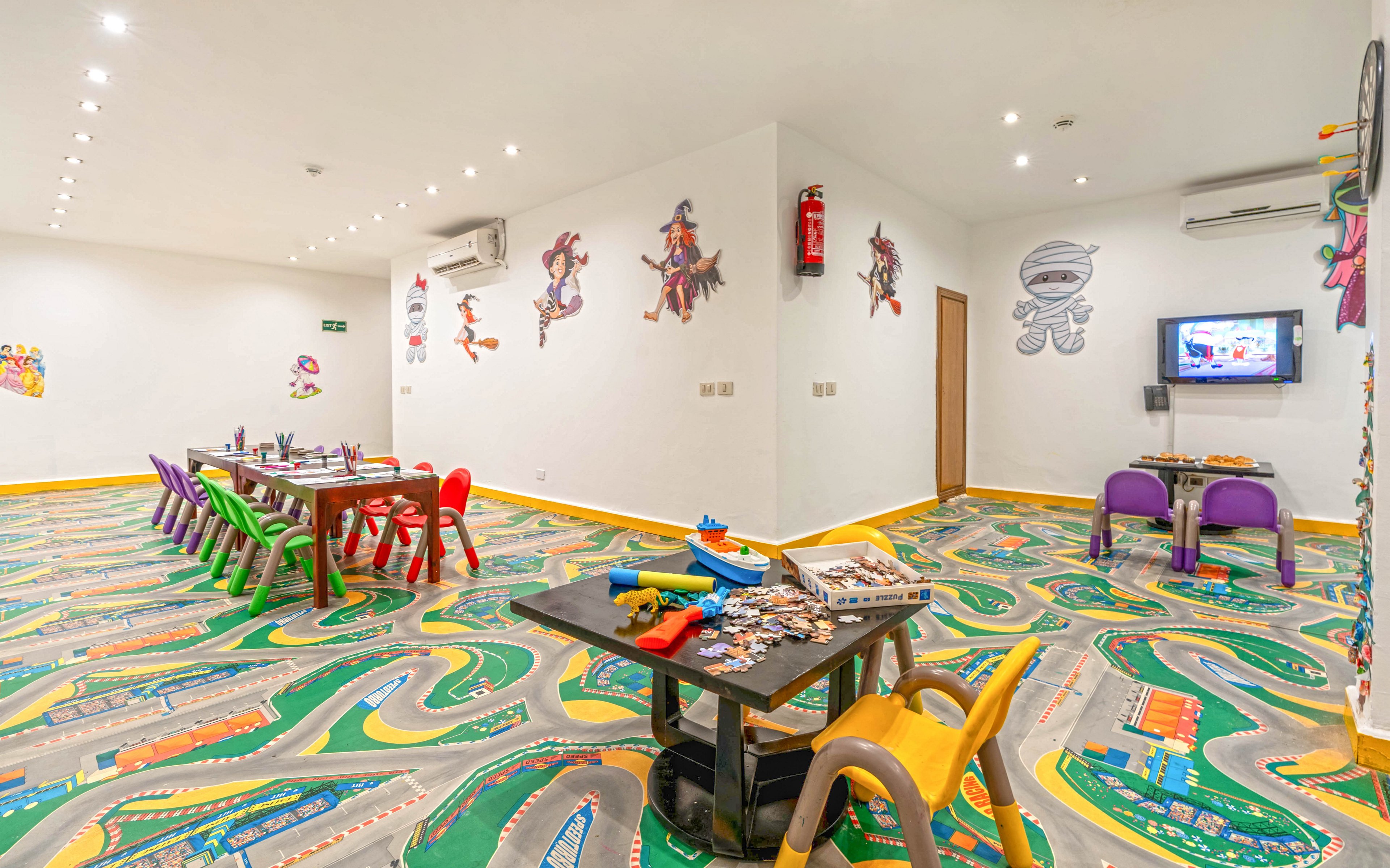 Brightly lit indoor playroom features several child-sized tables and colorful plastic chairs arranged for group activities. The floor is decorated with a playful racetrack pattern, adding movement and fun to the space. Walls display cartoon character decals, and a small television is mounted on one side of the room. Toys and craft materials are spread across the tables, creating an inviting environment for children.