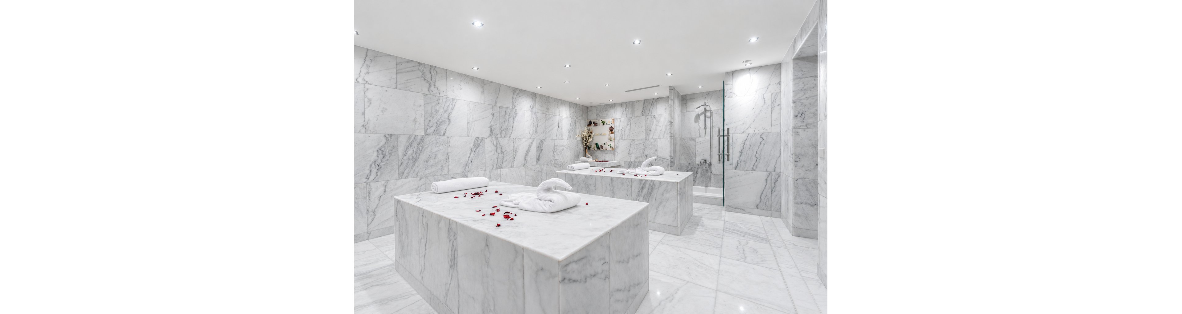 A bright, modern spa room features large marble massage tables topped with neatly rolled white towels. Red rose petals are scattered across the surfaces, adding a romantic accent to the cool stone. The space is fully clad in light gray veined marble, with recessed ceiling lights creating an airy, serene atmosphere. A glass door and subtle decorative elements enhance the clean, minimalist design.
